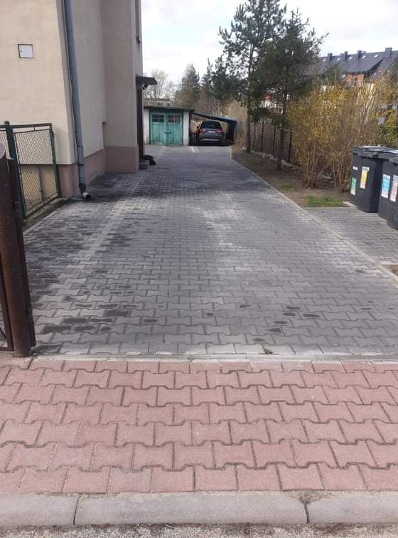 Długa, szara kostka brukowa prowadząca do garażu z zaparkowanym samochodem, obok budynku i rzędu czarnych koszy na śmieci.
