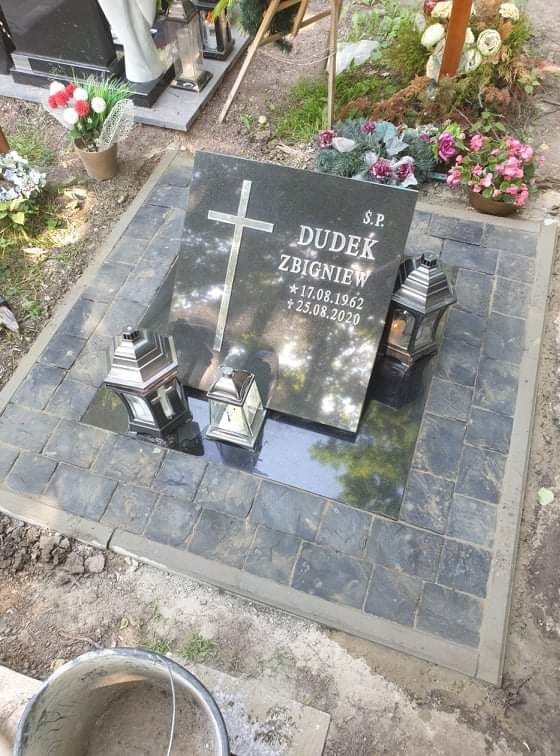 Czarny nagrobek z wygrawerowanym krzyżem i danymi osoby zmarłej, otoczony kostką brukową w odcieniach szarości i czerni, z lampionami na płycie nagrobnej i kwiatami w tle.