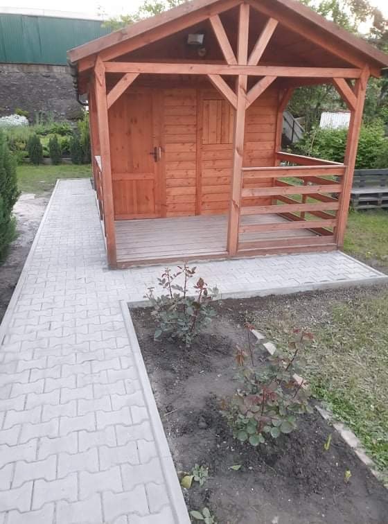 Drewniana altana z zamykanymi drzwiami, otoczona ścieżką z kostki brukowej w Gliwicach, z rabatą róż w pierwszym planie.