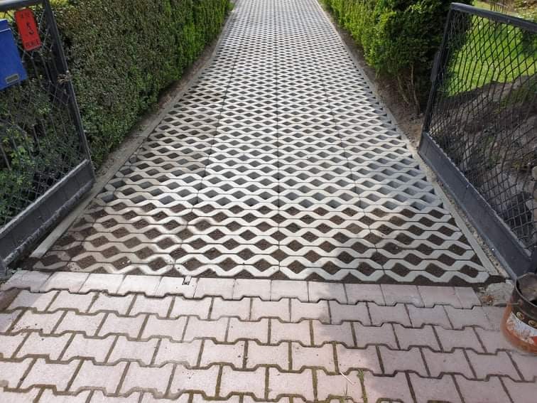 Podjazd z ażurowej kostki brukowej, widok z poziomu gruntu, otwarte metalowe furtki, po bokach wysokie, zielone krzewy.