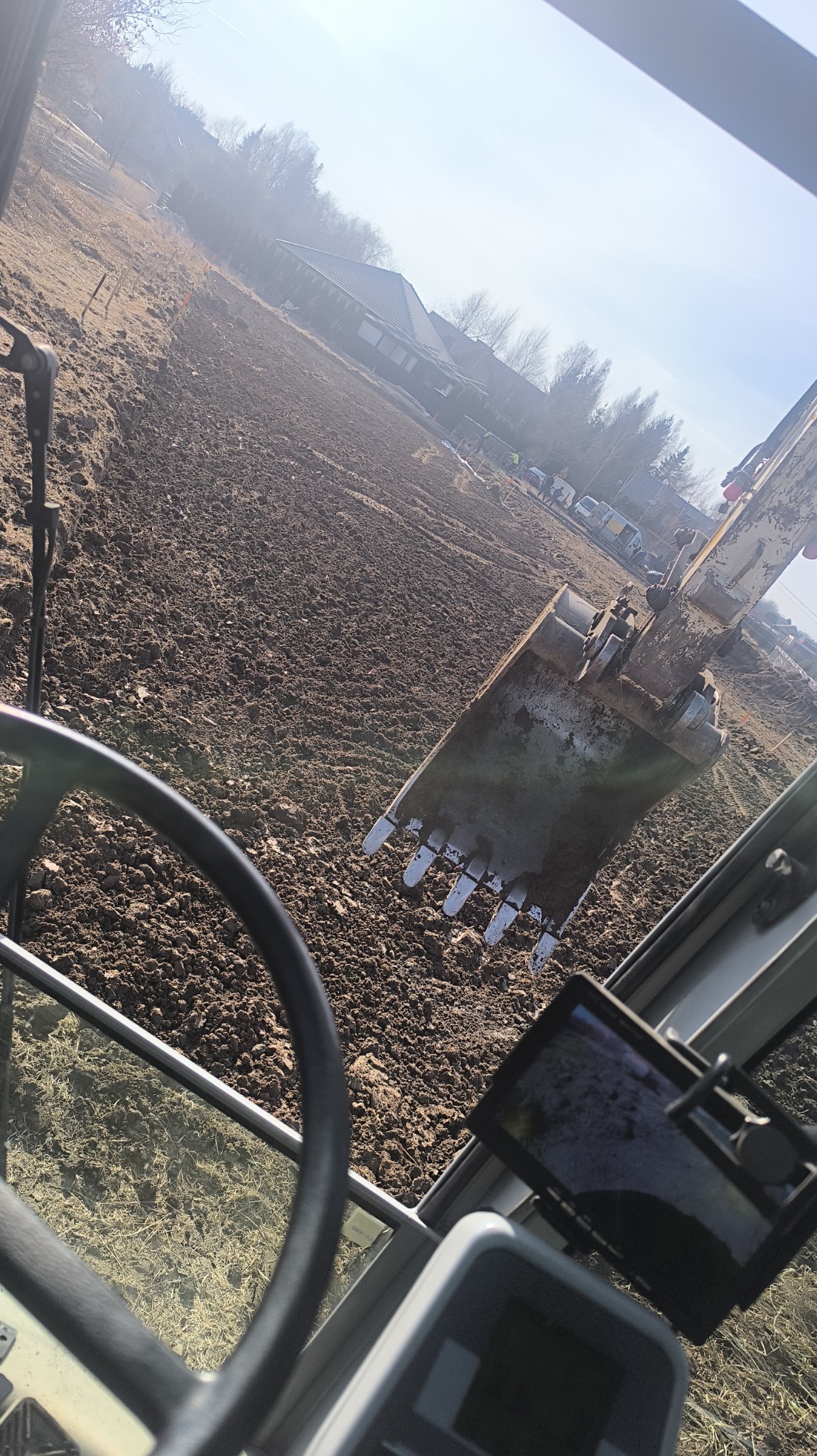 Widok z kabiny koparki na wyrównywany teren, widoczny lemiesz zagłębiający się w ziemię, w tle dom jednorodzinny i ekipa budowlana.