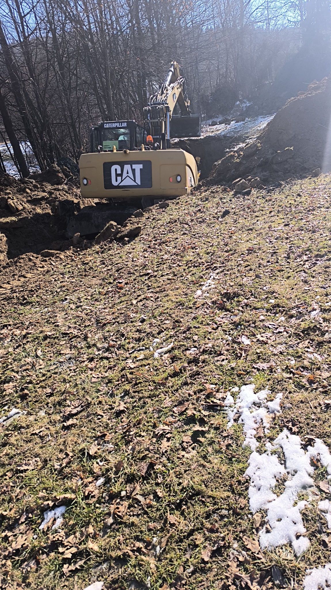 Żółta koparka Caterpillar w trakcie prac ziemnych, widoczny wykop w ziemi z resztkami śniegu, drzewa w tle.