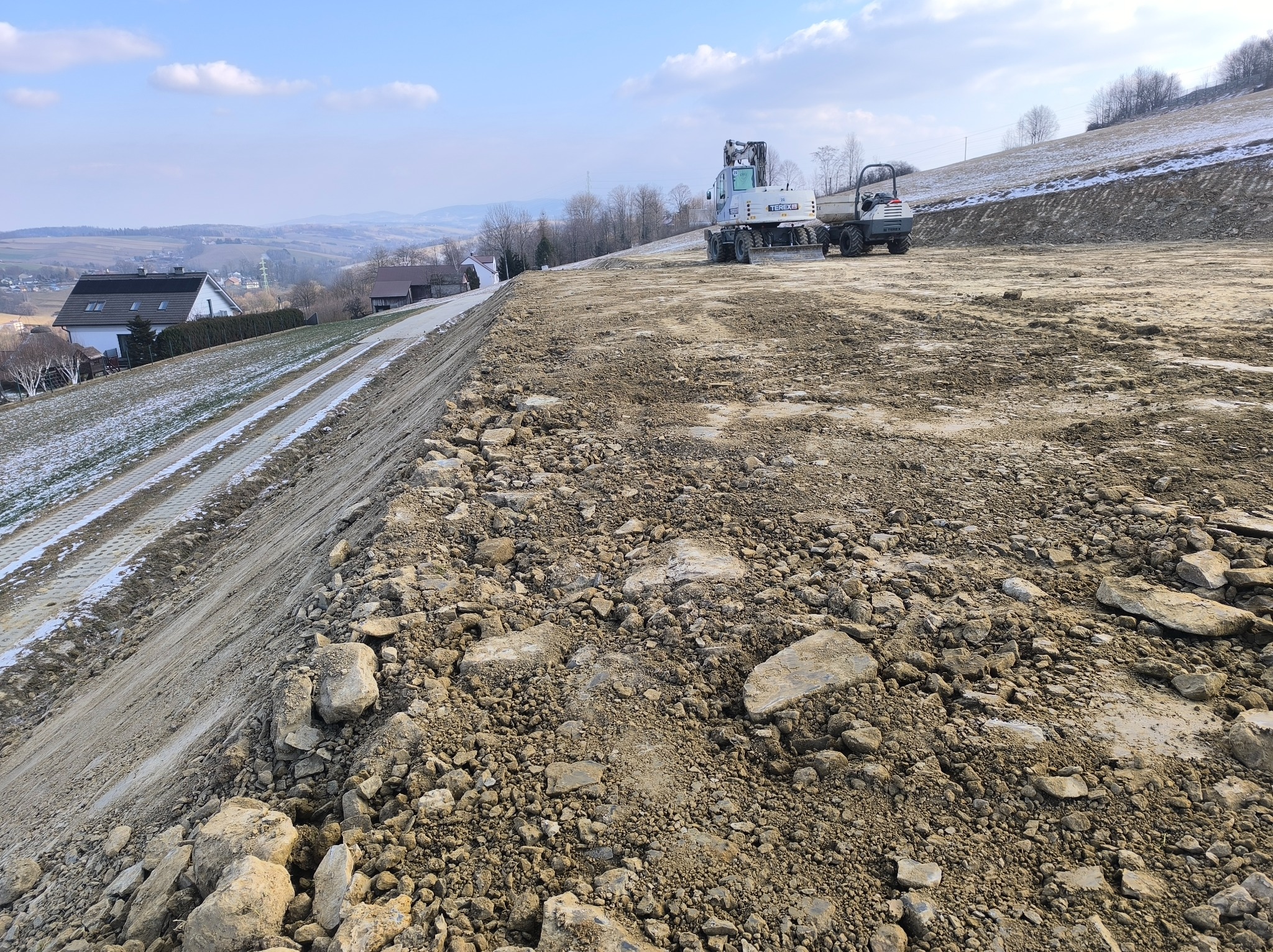 Wyrównany teren pod budowę na zboczu wzgórza, widoczny biały walec drogowy i koparka na tle zabudowań mieszkalnych i zalesionych wzniesień.