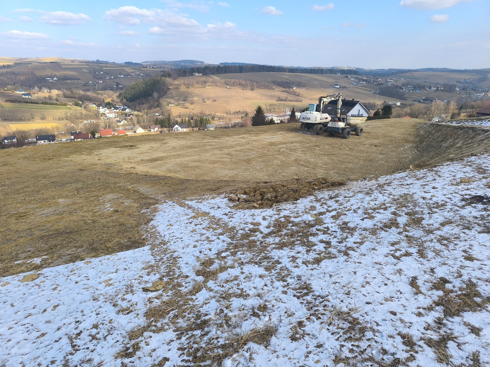 Dwie białe koparki Terex na wyrównanym terenie, w tle pagórkowaty krajobraz z zabudowaniami, resztki śniegu na pierwszym planie.