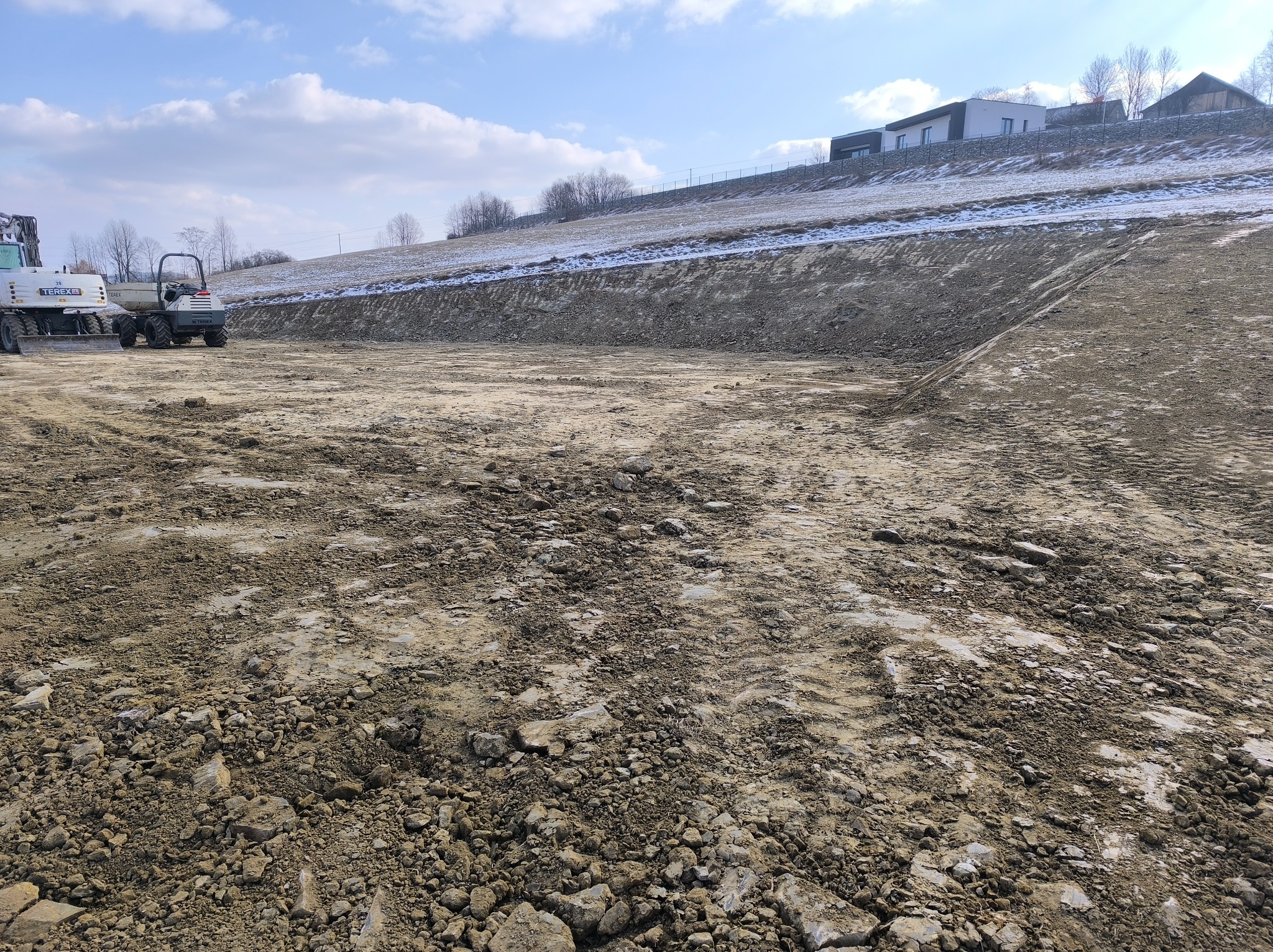 Przygotowany teren pod budowę, z widocznymi śladami opon maszyn budowlanych, w tle skarpa z resztkami śniegu i budynki mieszkalne pod błękitnym niebem.