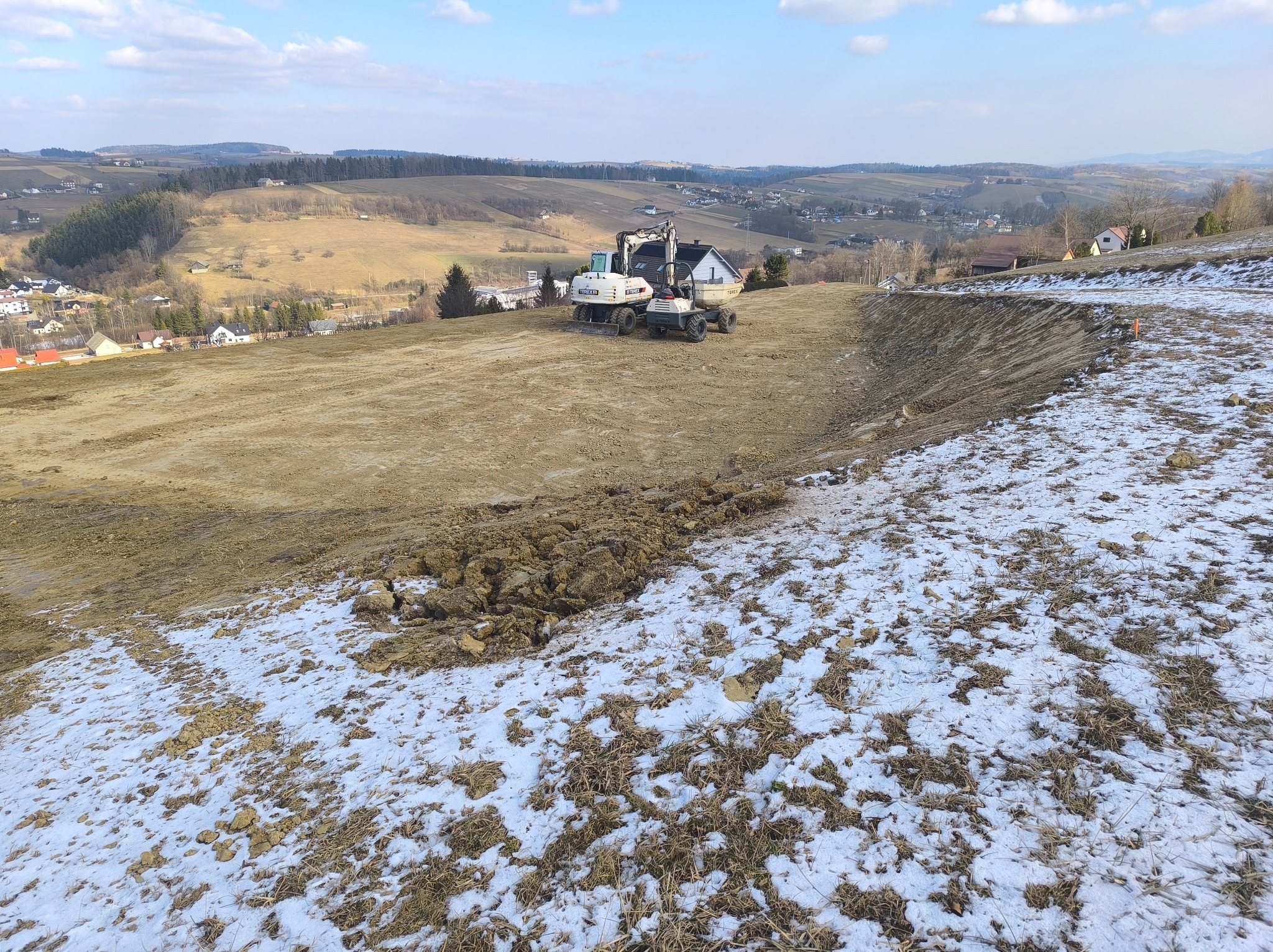 Dwa białe pojazdy budowlane, w tym koparka, na wyrównanym terenie z resztkami śniegu i widokiem na pagórkowaty krajobraz w tle.