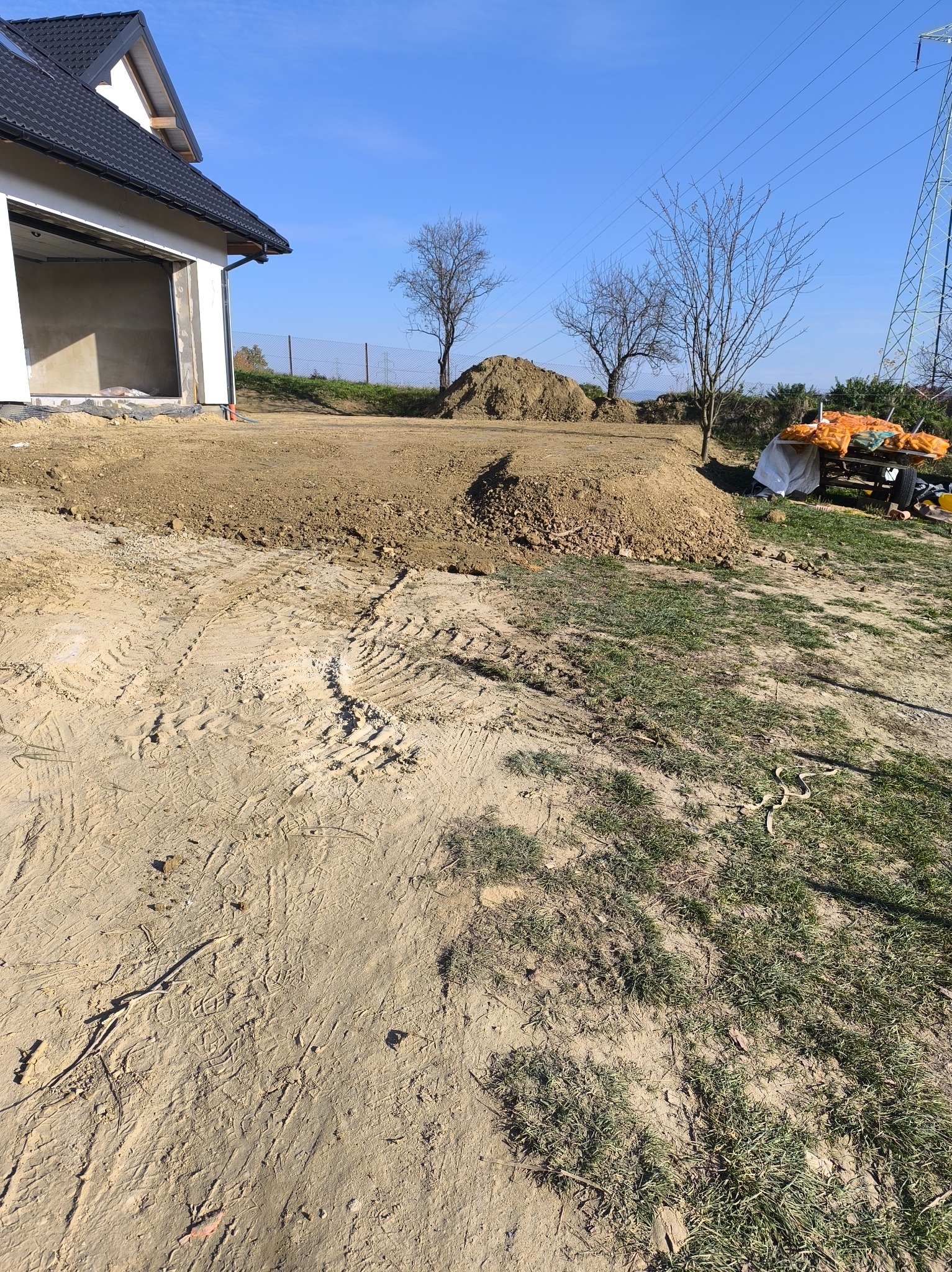 Teren budowy z widocznymi śladami opon, świeżo usypana ziemia, przygotowana pod budowę domu jednorodzinnego z garażem w stanie surowym otwartym, słoneczny dzień.