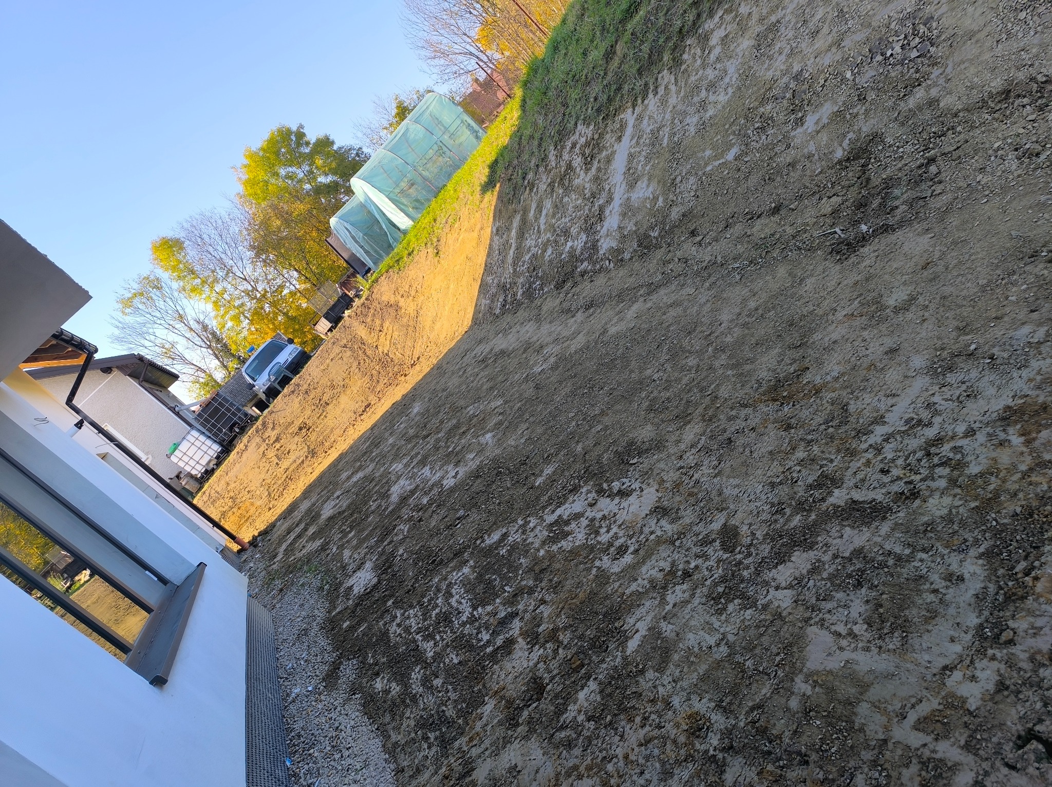 Przygotowany teren pod budowę z widoczną ścianą wykopu, zielenią w tle i fragmentem budynku z oknami.