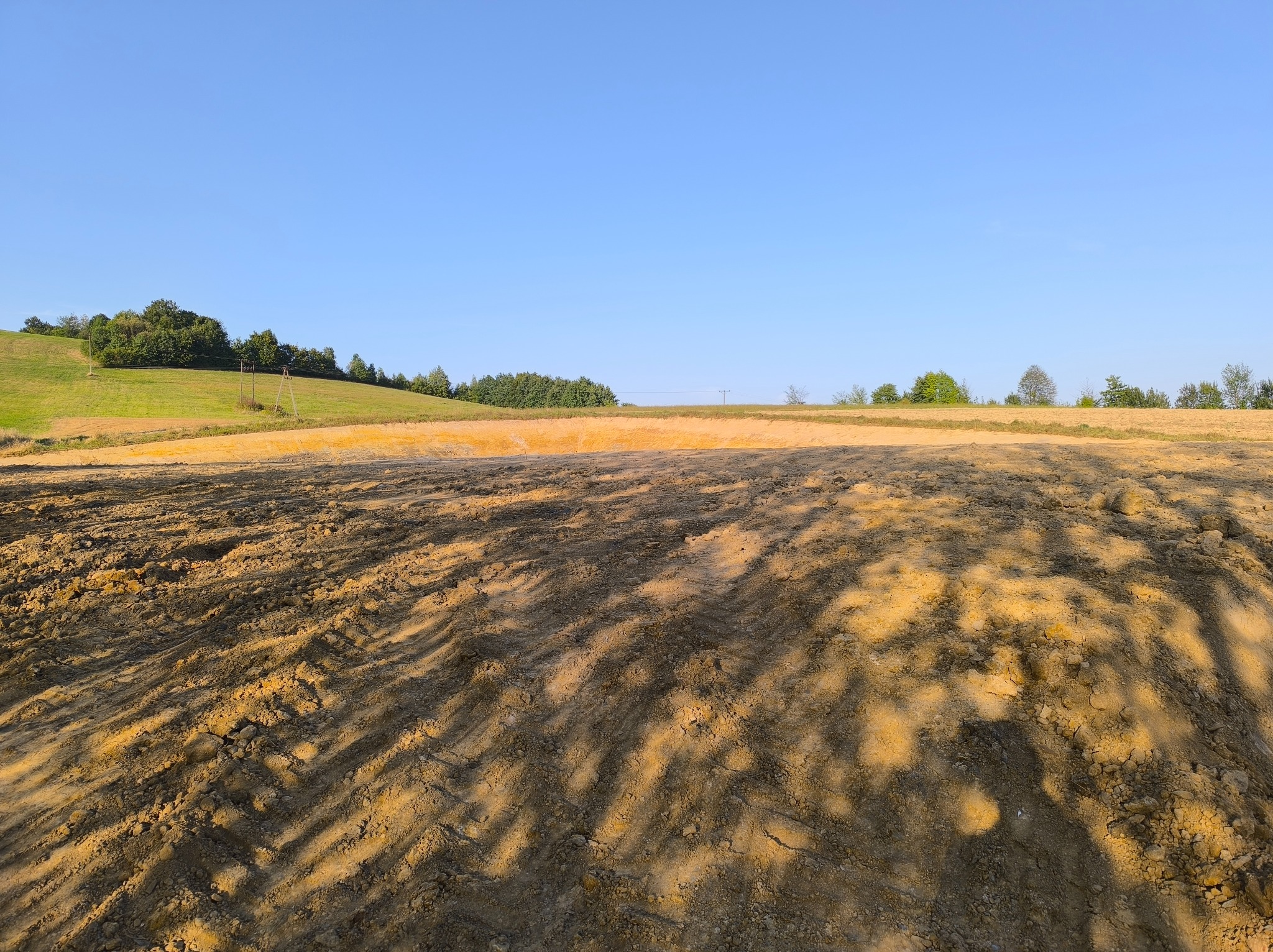 Świeżo wyrównany teren pod budowę z widocznymi śladami po maszynach, w tle łagodny zielony pagórek i błękitne niebo.