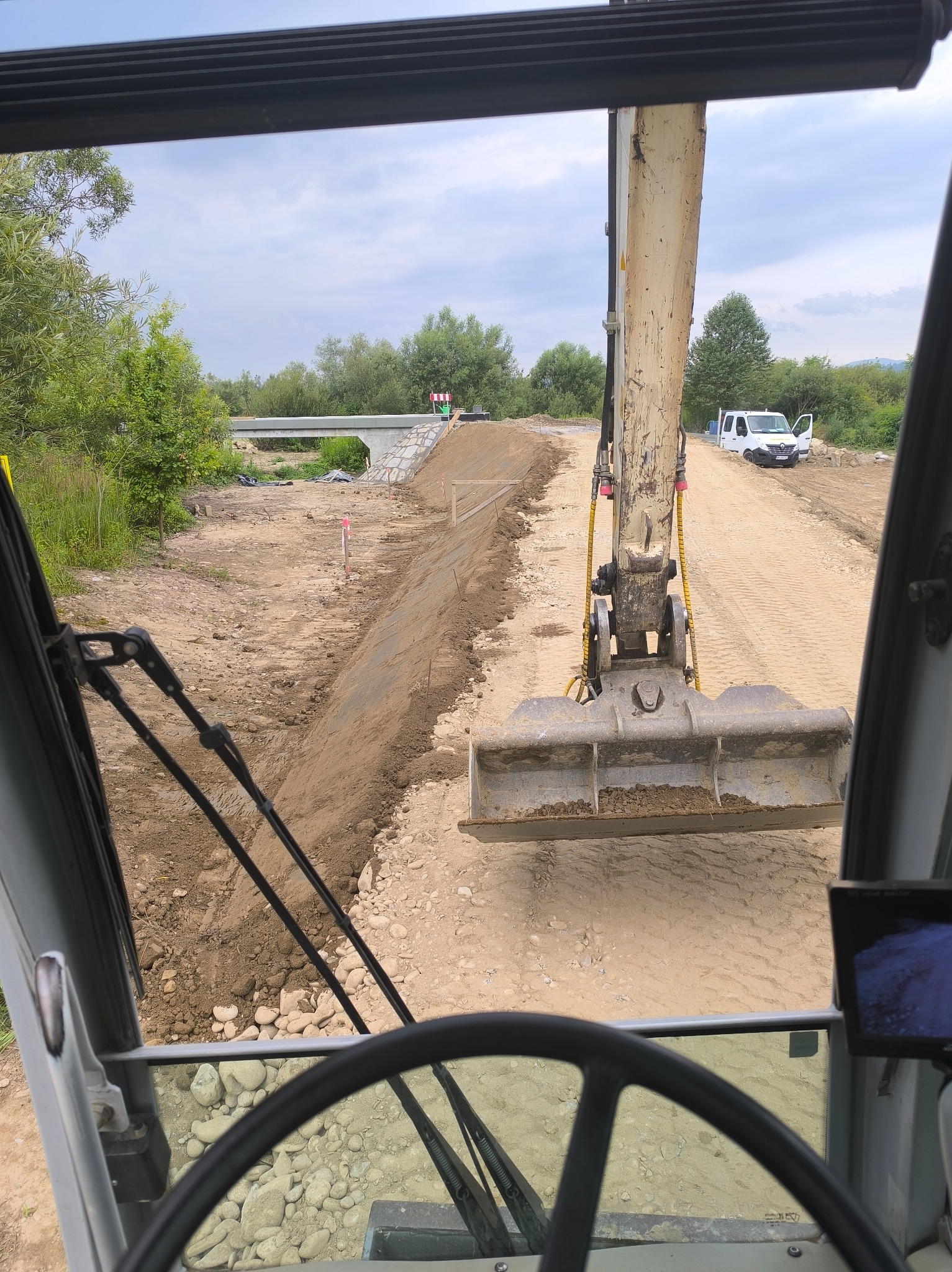 Widok z kabiny koparki na plac budowy z wyrównanym terenem, w tle most i biały samochód dostawczy.
