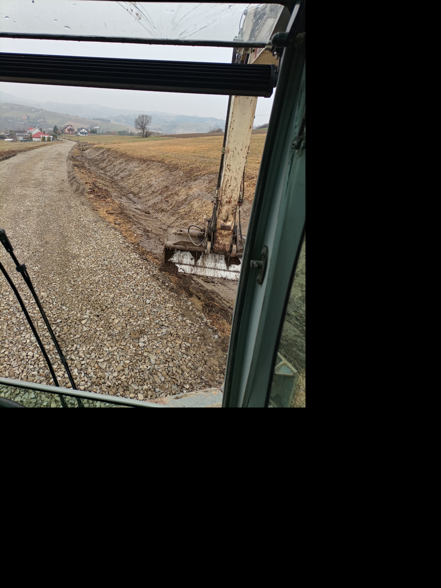 Widok z kabiny koparki na rów wykopany w ziemi, obok droga żwirowa, w tle zabudowania wiejskie w pochmurny dzień.