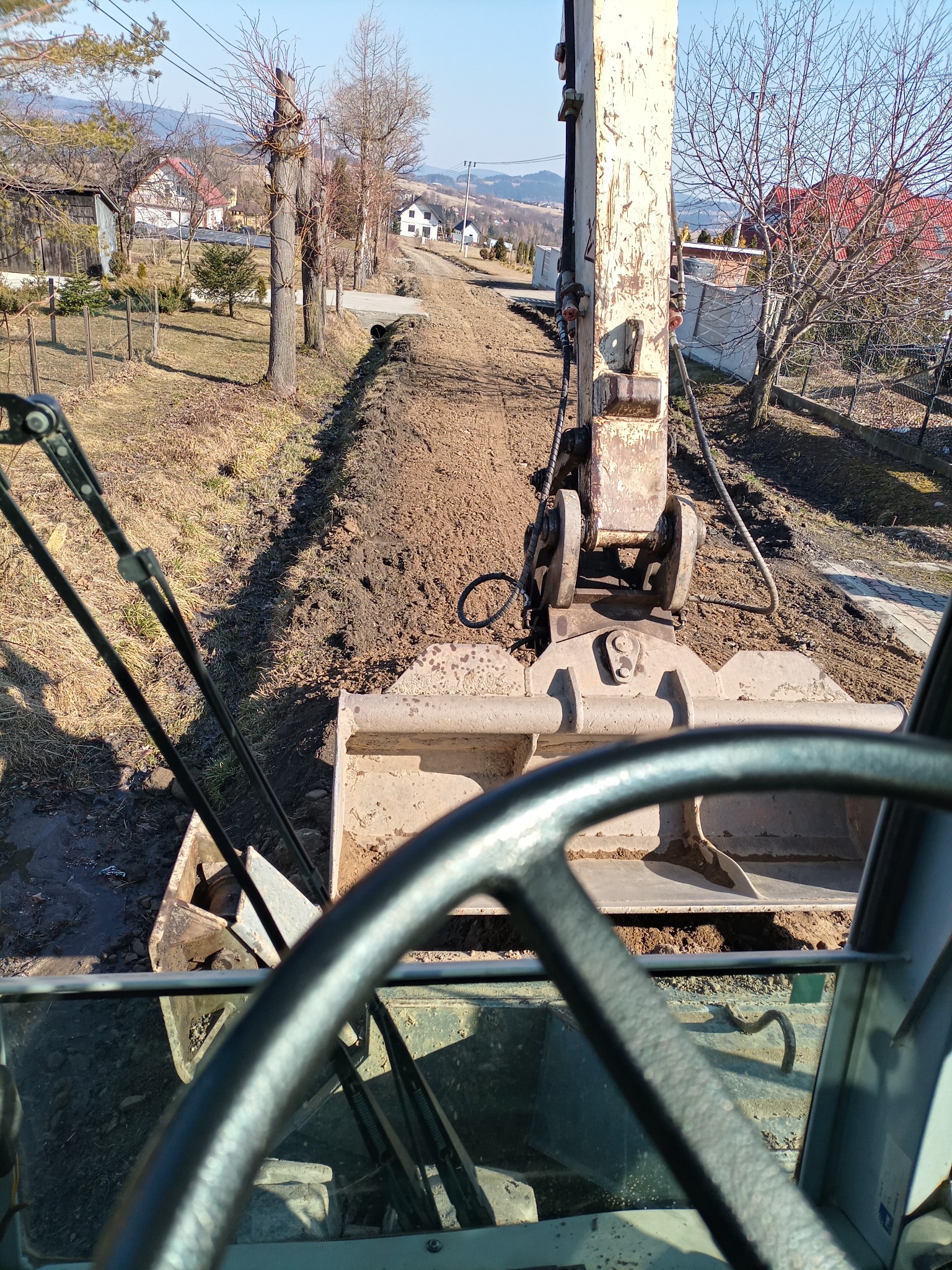Widok z kabiny koparki na wykopany rów w ziemi, w tle zabudowania wiejskie i wzniesienia.
