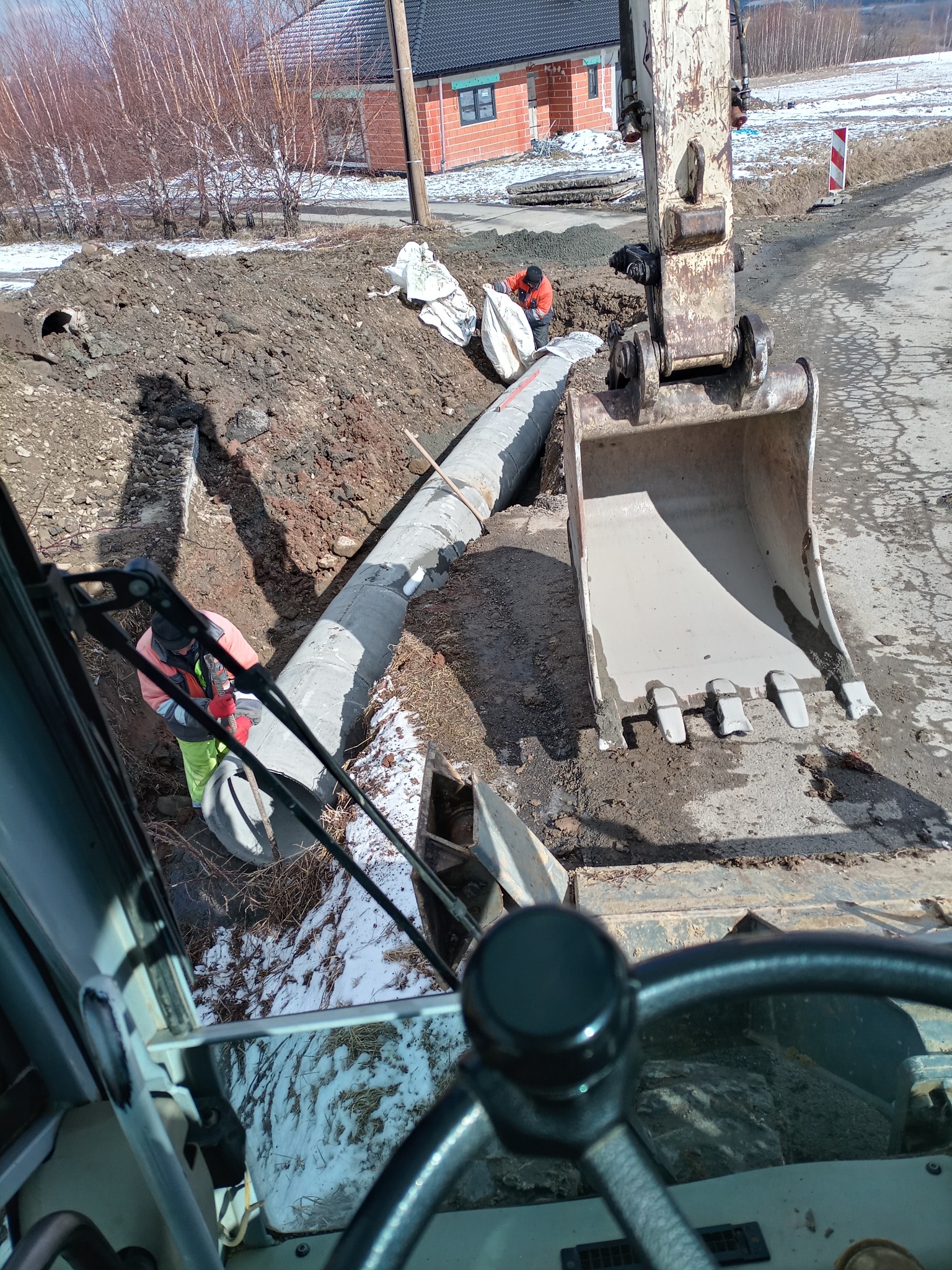 Widok z kabiny koparki na robotników układających betonową rurę w wykopie, w tle częściowo ośnieżony teren i budynek w trakcie budowy.