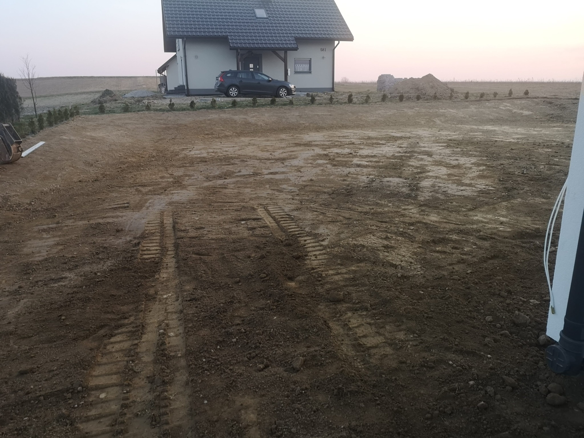 Przygotowany teren pod budowę domu jednorodzinnego, widoczne ślady gąsienic, wyrównana ziemia, w tle dom z samochodem i nasadzenia z małych drzewek.