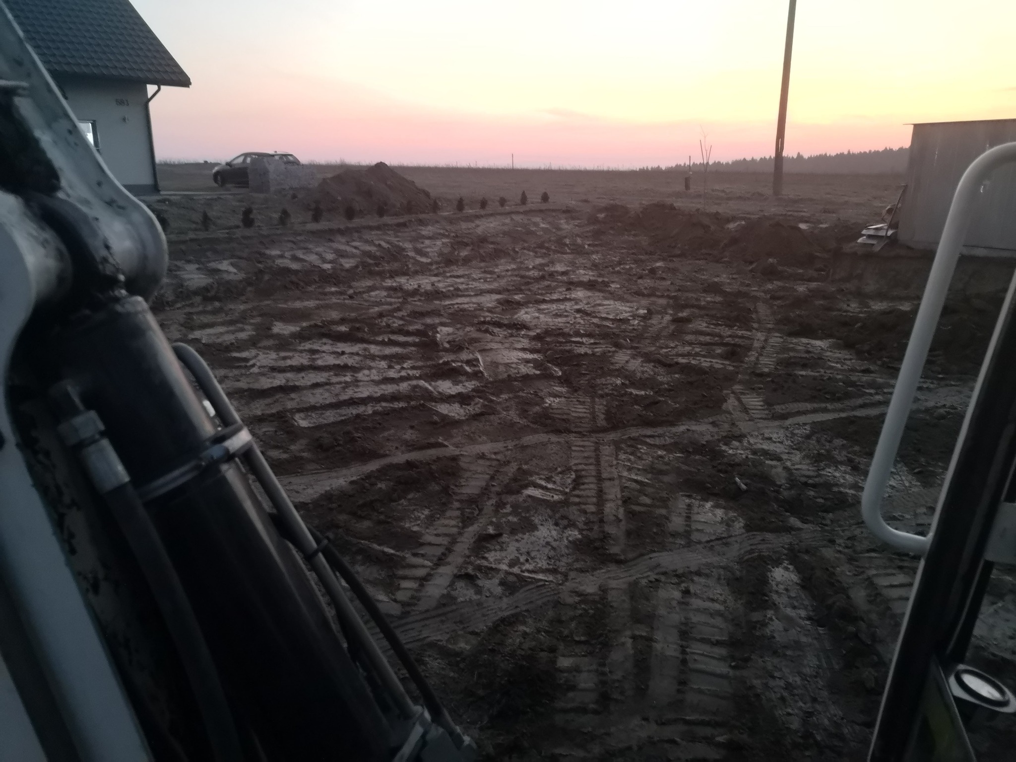 Widok z kabiny maszyny budowlanej na teren budowy o zmierzchu, widoczne ślady opon na błotnistym podłożu, w tle wzniesiona ziemia i fundamenty domu.