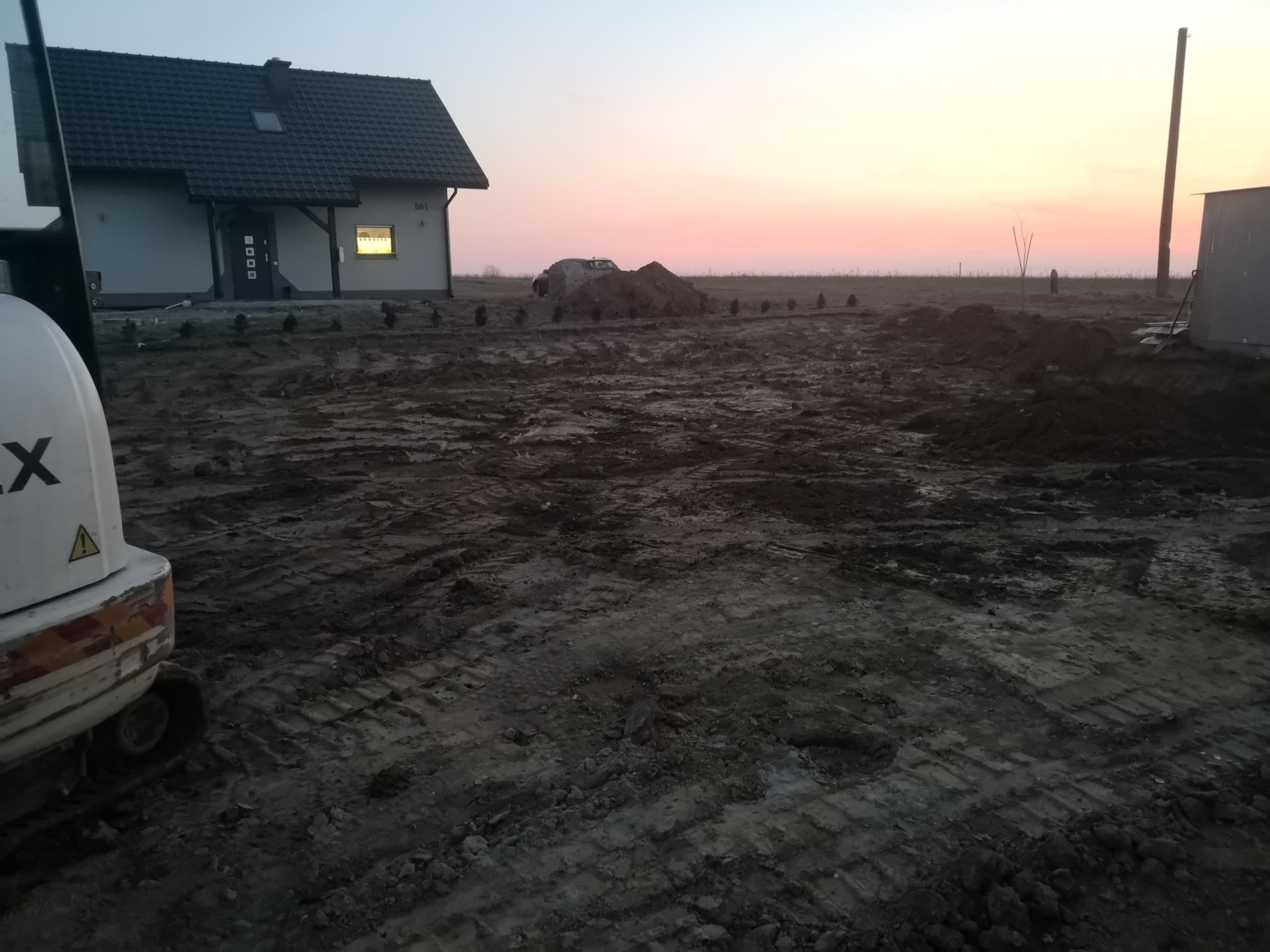 Przygotowany teren pod budowę domu jednorodzinnego podczas zachodu słońca, widoczne ślady opon po maszynach budowlanych, małe drzewka wzdłuż granicy działki i mini koparka z lewej strony.