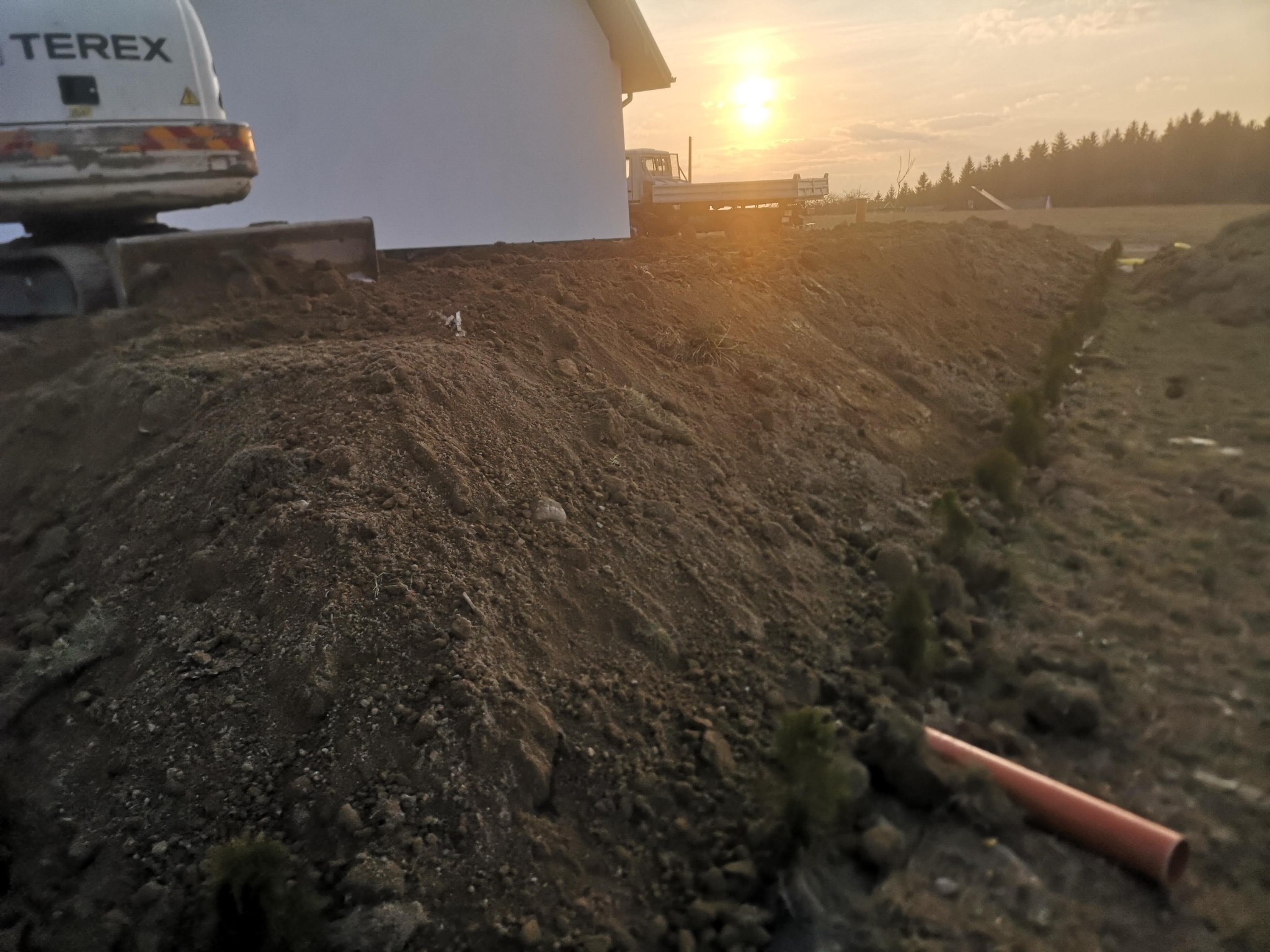 Częściowo wykopany rów w ziemi z widocznym fragmentem koparki Terex z tyłu po lewej, rurą PCV na pierwszym planie i młodymi drzewkami po prawej stronie, w tle budynek i zachodzące słońce.