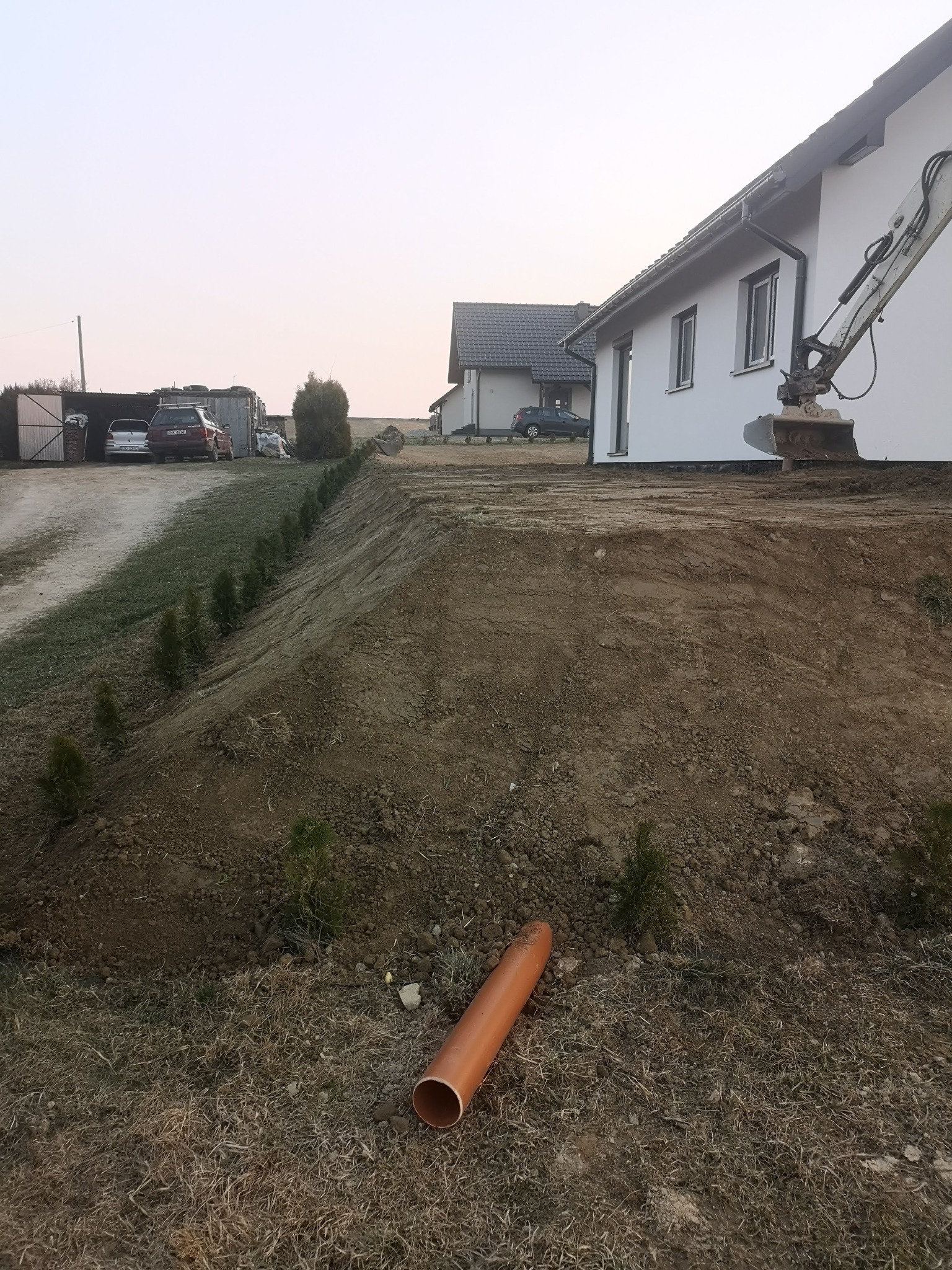 Przygotowany teren pod budowę domu z widocznym wykopem, rzędem małych drzewek iglastych i pomarańczową rurą kanalizacyjną leżącą na trawie, w tle domy jednorodzinne i koparka.
