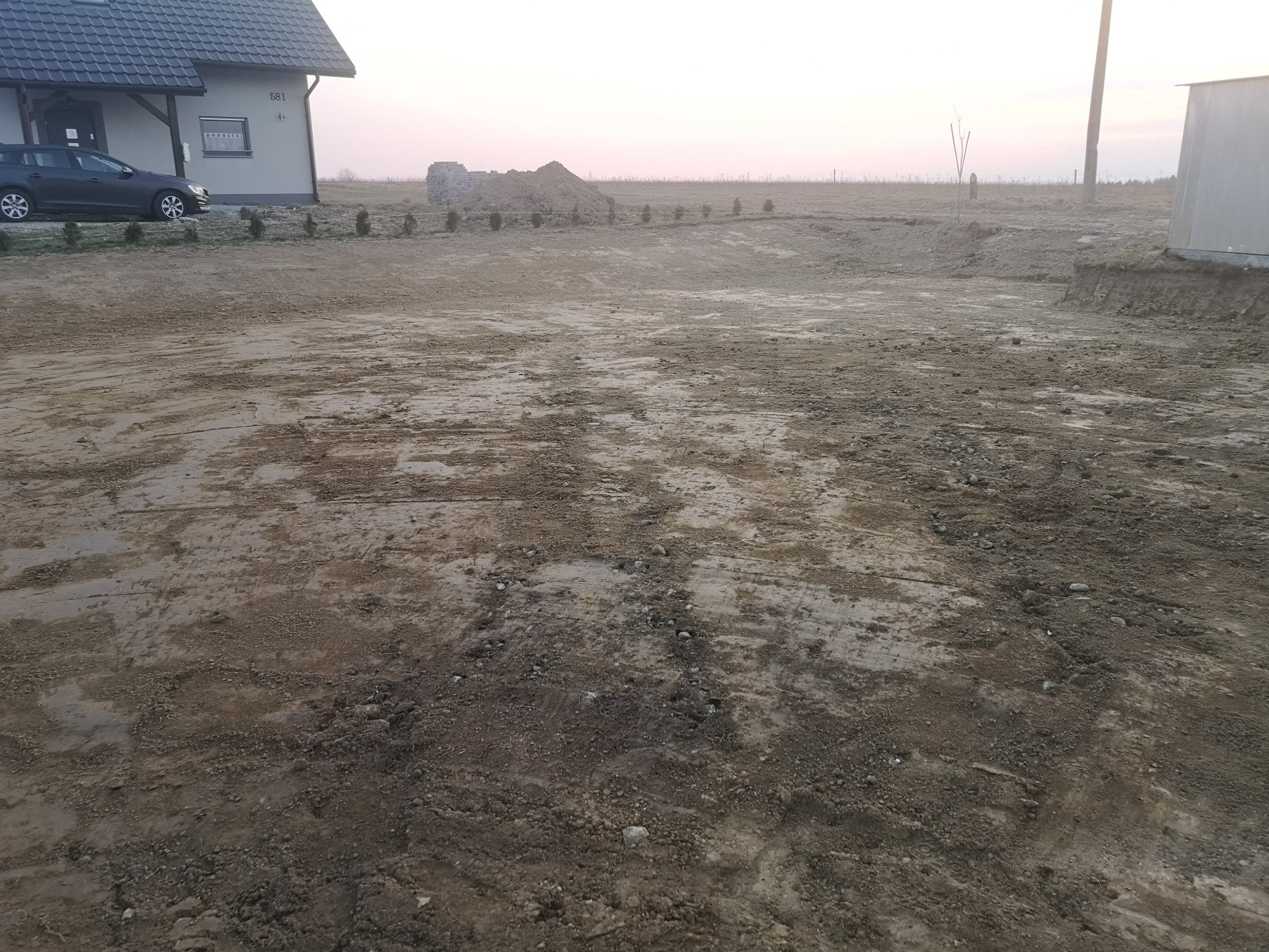 Przygotowany teren pod budowę domu, z wyplantowaną ziemią, widoczne ślady opon i niewielkie drzewka wzdłuż granicy działki, w tle sterta ziemi i częściowo wykończony budynek.