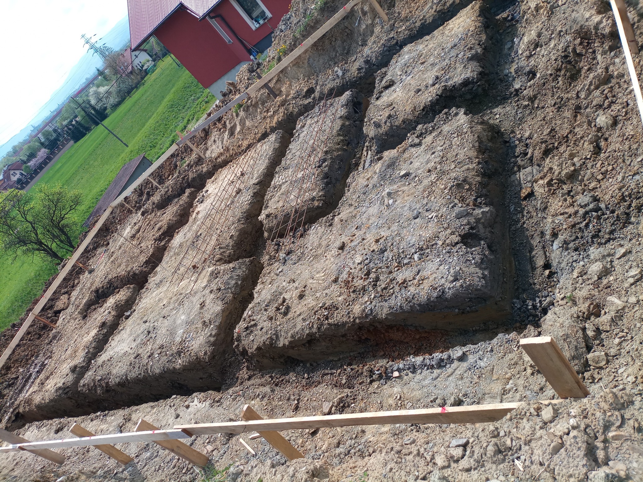 Wykopane rowy pod fundamenty z widocznym zbrojeniem, obudowane drewnianymi deskami, na tle zieleni i czerwonego domu.