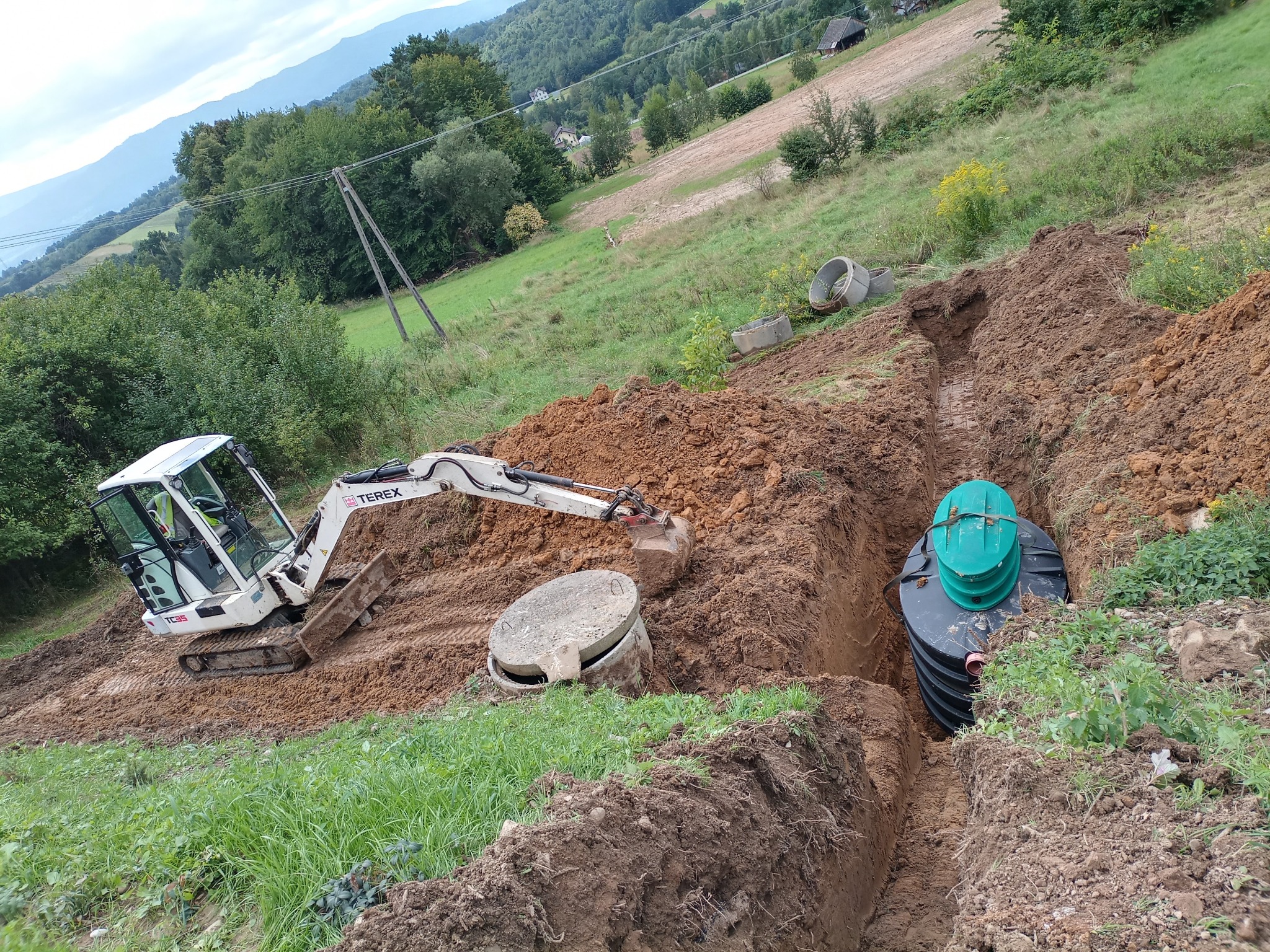Mała koparka Terex obok wykopu z wstawionym zbiornikiem z zieloną pokrywą na tle pagórkowatego krajobrazu.