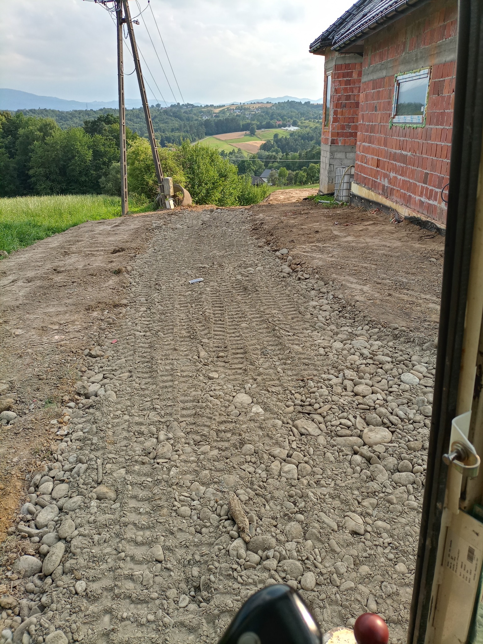 Widok z kabiny maszyny budowlanej na teren przygotowany pod budowę domu jednorodzinnego, wyrównany i wysypany kamieniami, w tle malowniczy krajobraz zalesionych wzgórz.