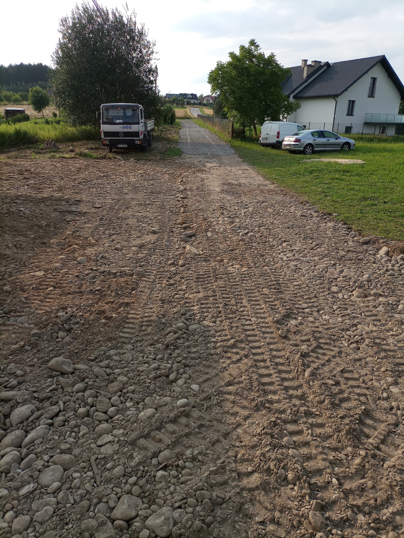 Udrożniona droga gruntowa z licznymi śladami opon, widoczny samochód ciężarowy na początku drogi oraz dom jednorodzinny z tyłu.
