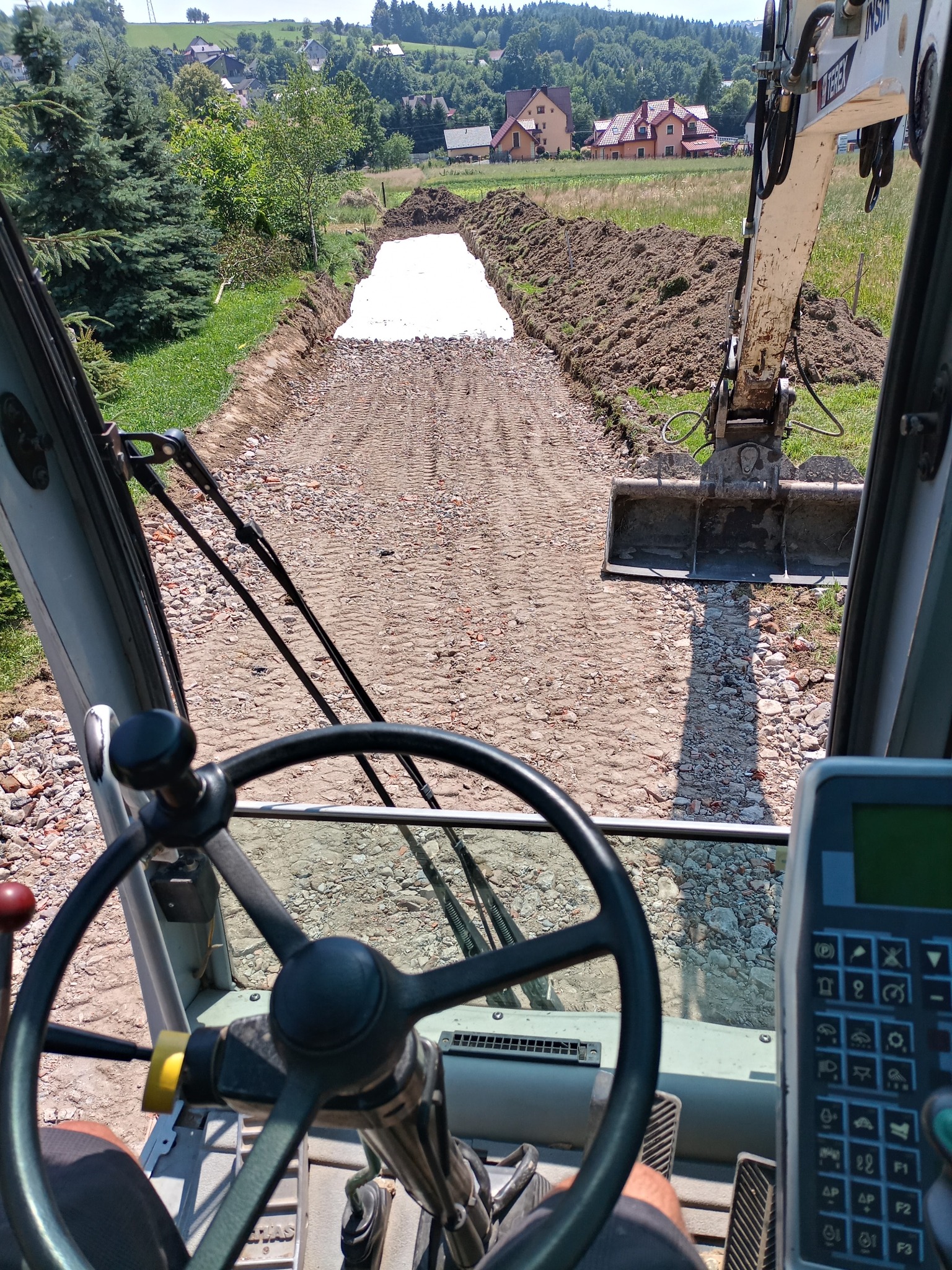 Widok z kabiny operatora koparki na wykop pod fundamenty, z widocznym białym geowłókniną na dnie wykopu, w tle zabudowa wiejska i zalesione wzgórza.