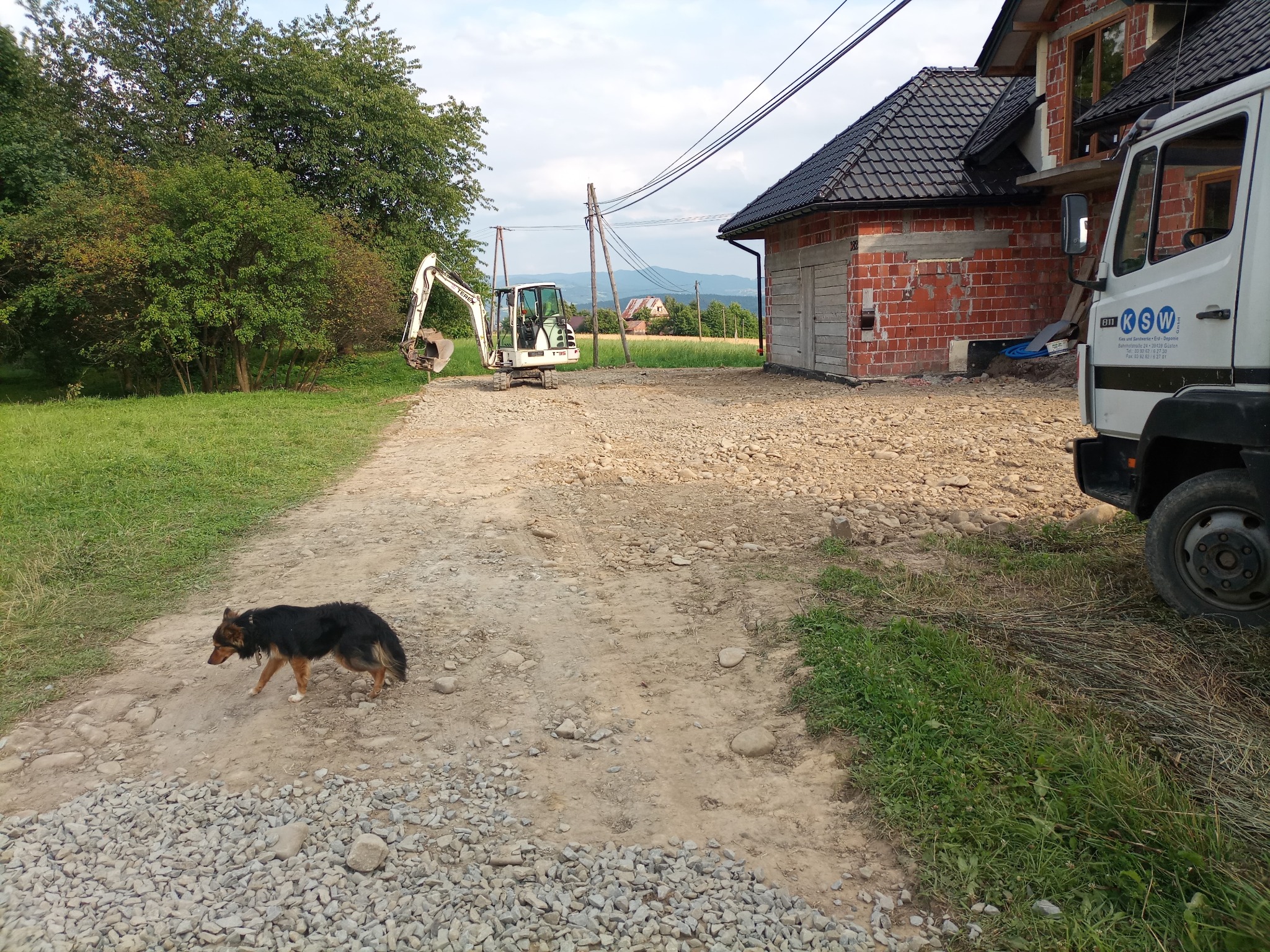 Biała mini-koparka Terex T35 na żwirowej drodze przed budynkiem w trakcie budowy, w tle krajobraz z widocznymi wzniesieniami, na pierwszym planie pies rasy mieszanej.
