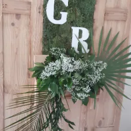 Dekoracja ślubna z drewnianego panelu z mchu, białych liter G i R oraz kompozycją z zielonych liści i drobnych białych kwiatów.
