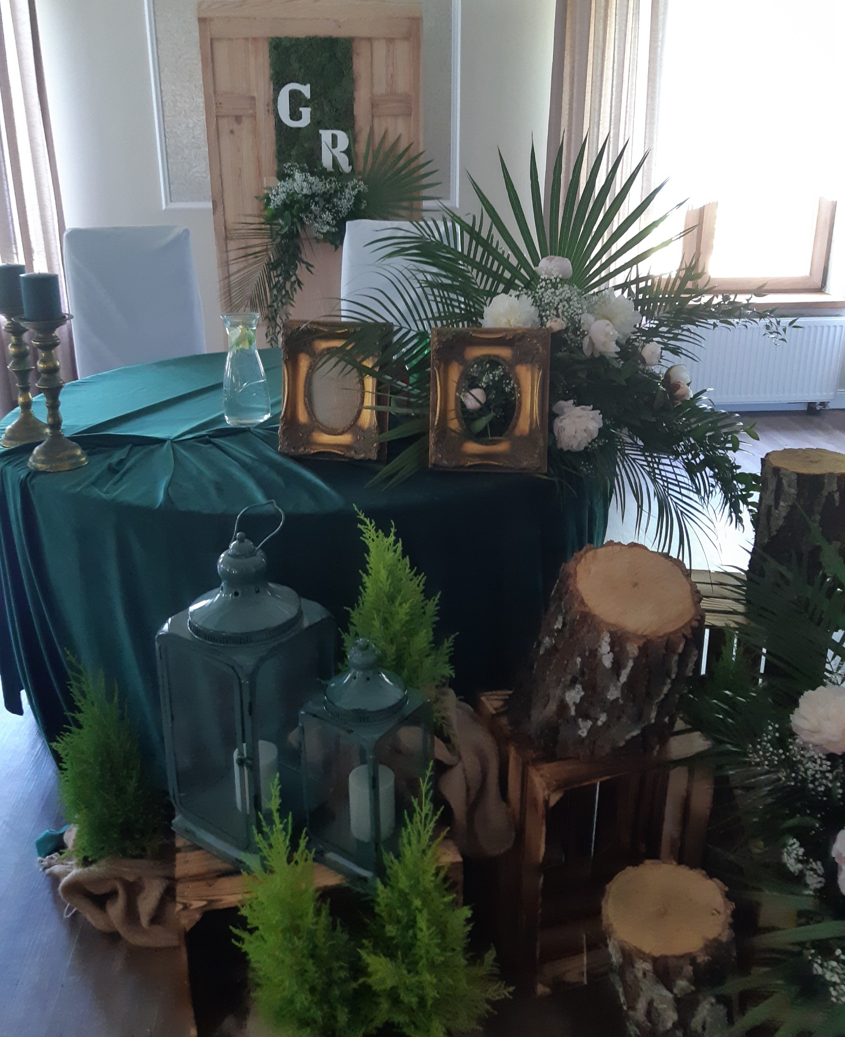 Dekoracja stołu weselnego z zielonym obrusem, lampionami, drewnianymi elementami i kwiatami, w tle drewniane drzwi z mchem i inicjałami GR.