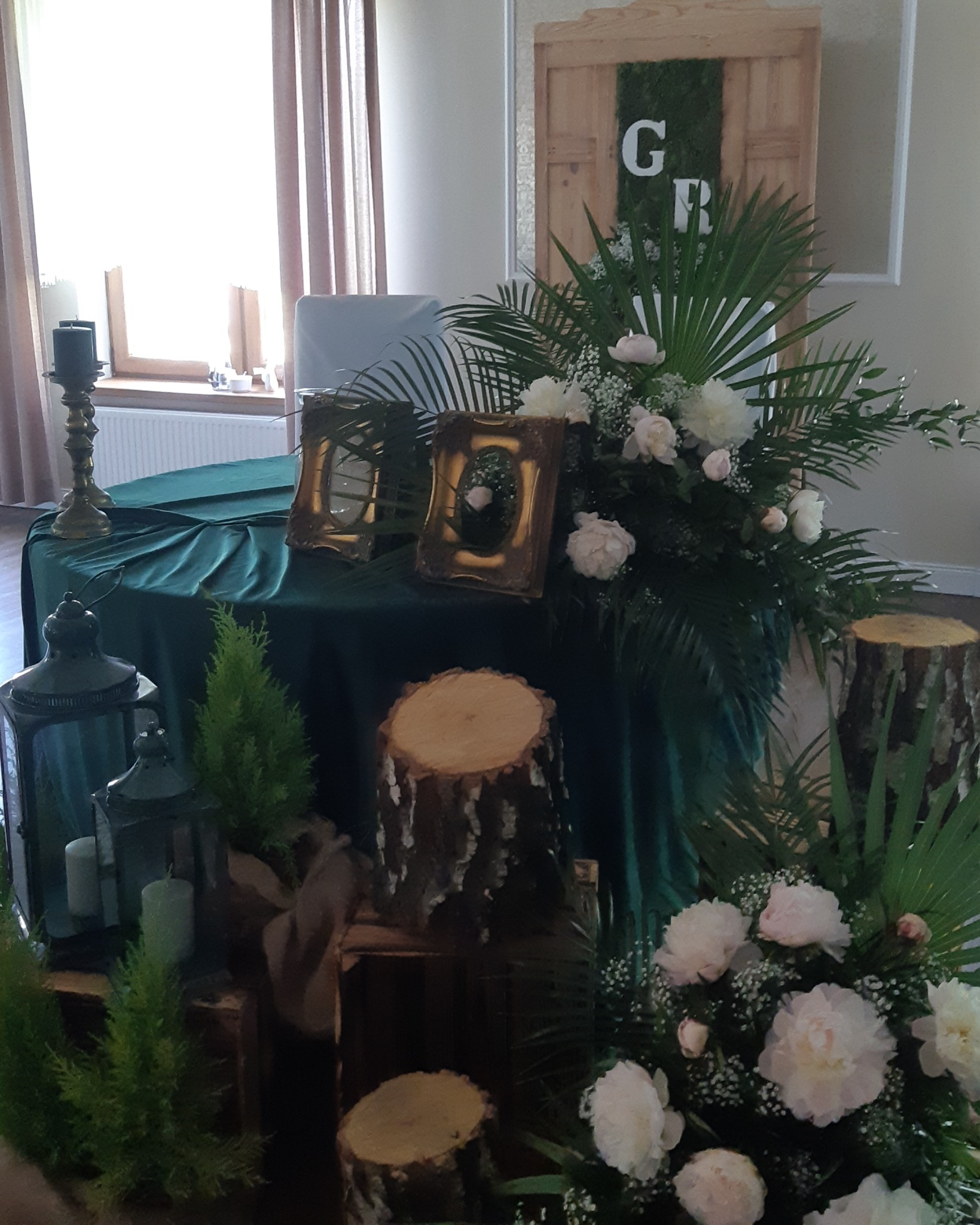 Dekoracja stołu na uroczystość z zielonym obrusem, drewnianymi elementami, świecami w lampionach i kompozycją kwiatową z białych piwonii i palm, z inicjałami 'G' i 'R' na drewnianym tle.