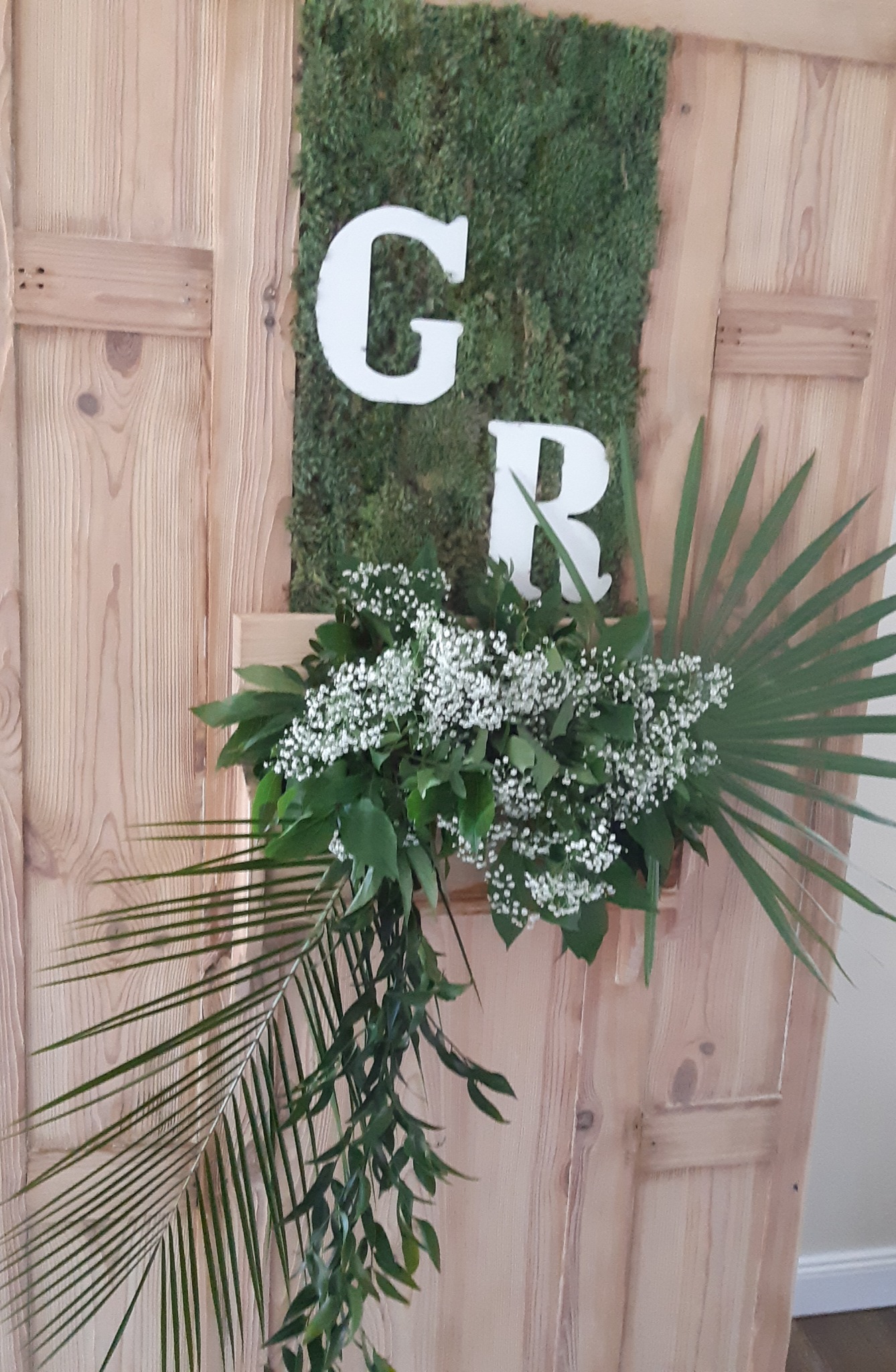Dekoracja ślubna z drewnianego panelu z mchu, białych liter G i R oraz kompozycją z zielonych liści i drobnych białych kwiatów.