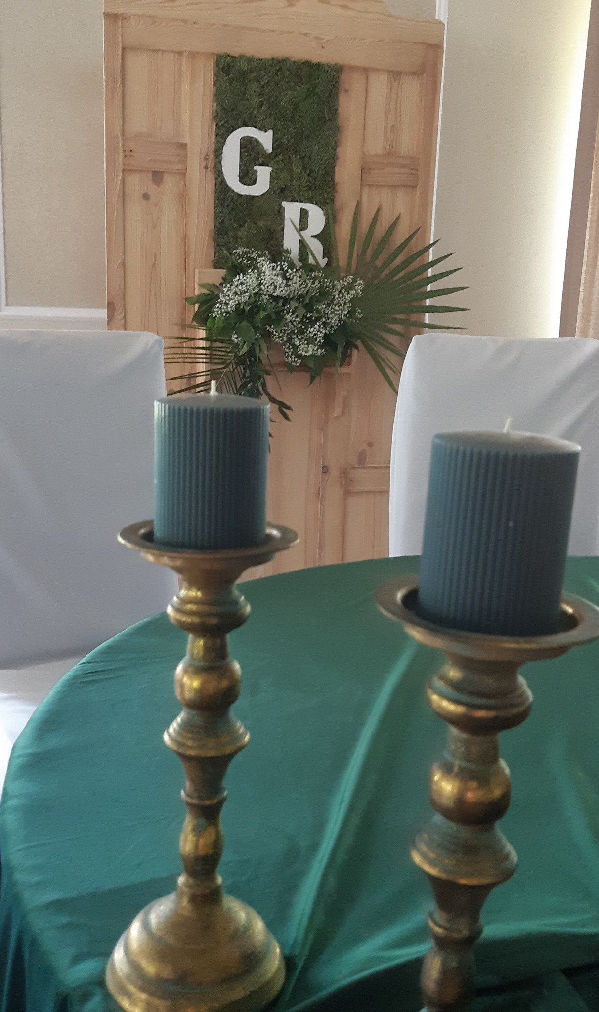 Dekoracja stołu weselnego: dwie szare świece w złotych świecznikach na zielonym obrusie, w tle drewniane drzwi z mchem i inicjałami 'G' i 'R' oraz kompozycją kwiatową.