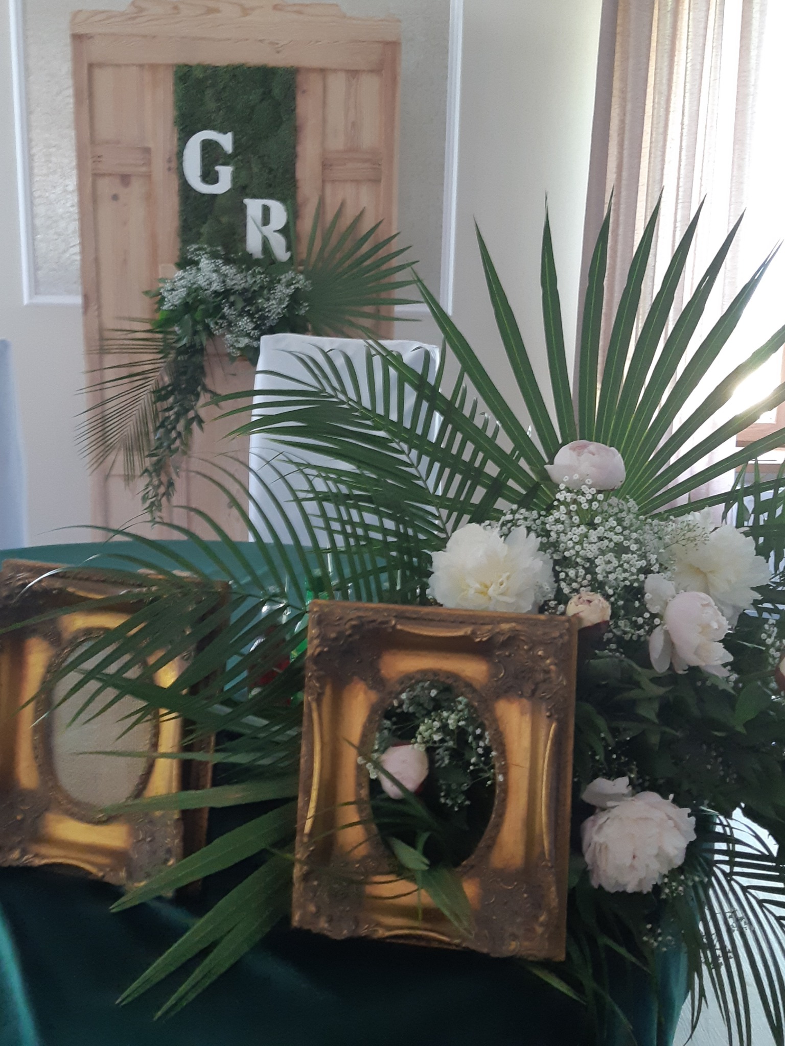 Dekoracja stołu weselnego z białymi piwoniami, gipsówką, liśćmi palmowymi i złotymi ramkami na zdjęcia, na tle drewnianej ściany z mchem i inicjałami 'G' i 'R'.