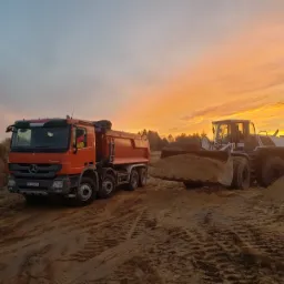 Pomarańczowa wywrotka Mercedes i biała ładowarka Liebherr z pełną łyżką piasku na tle pomarańczowego nieba o świcie w Skarżysku-Kamiennej.