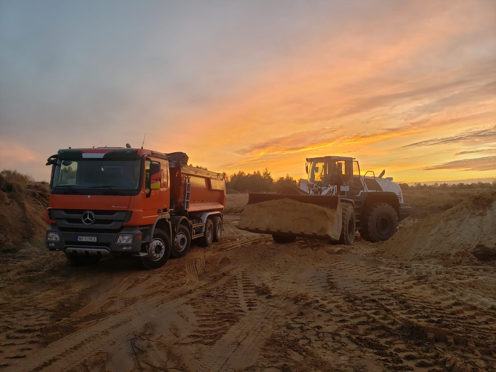 Pomarańczowa wywrotka Mercedes obok białej ładowarki z pełną łyżką urobku na tle wschodzącego słońca.