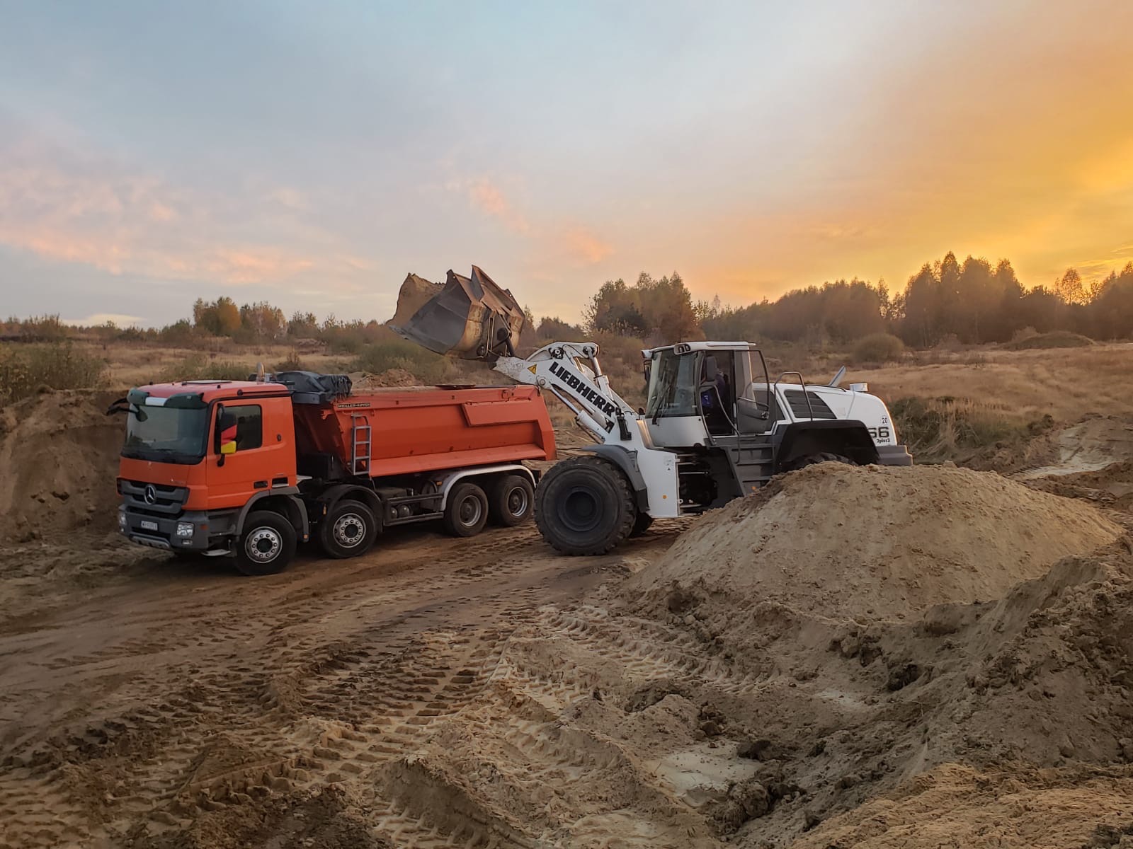 Pomarańczowa wywrotka Mercedes i biała ładowarka Liebherr podczas załadunku ziemi na placu budowy o zmierzchu.