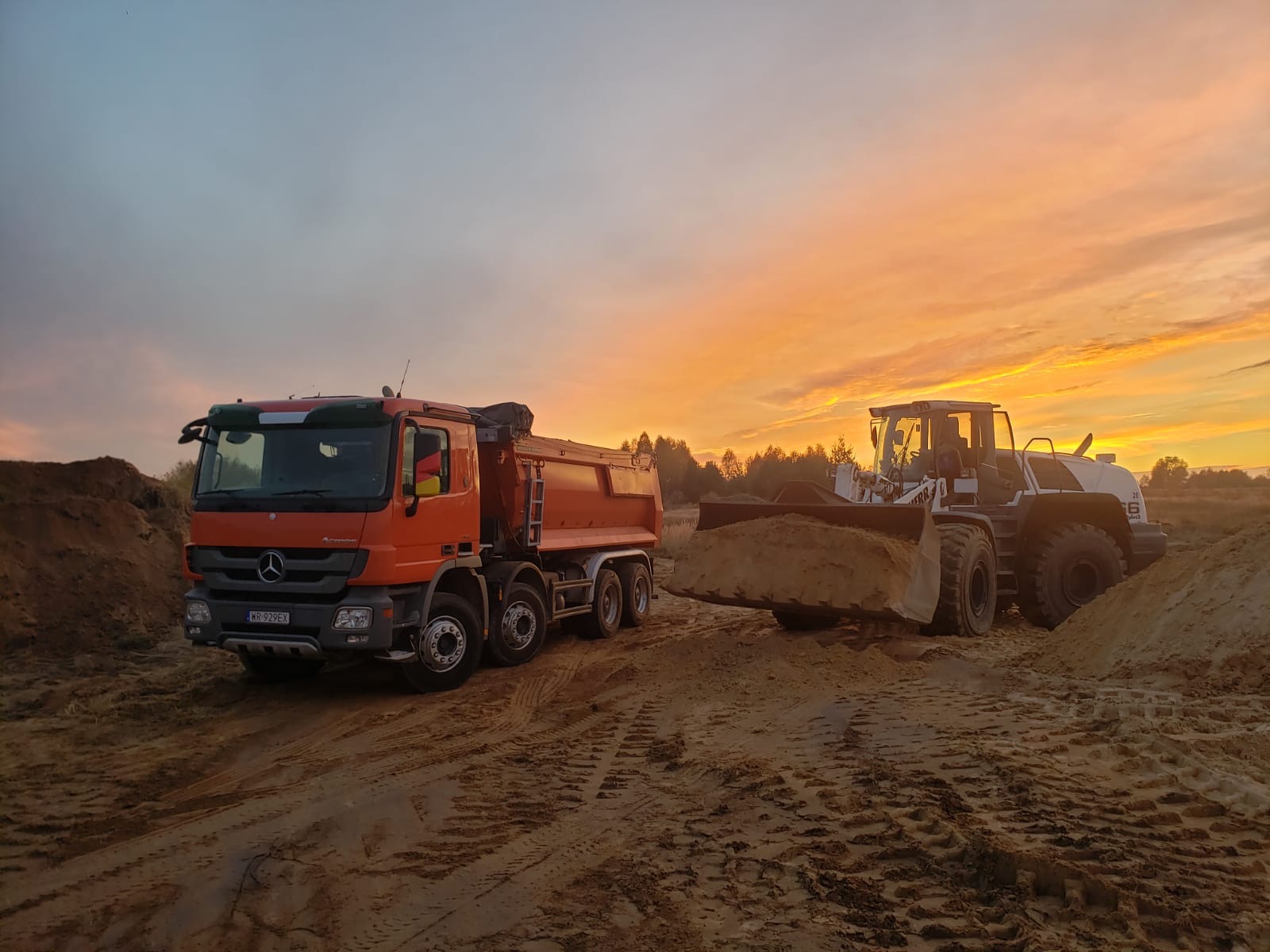 Pomarańczowa wywrotka Mercedes i biała ładowarka Liebherr z pełną łyżką piasku na tle pomarańczowego nieba o świcie w Skarżysku-Kamiennej.