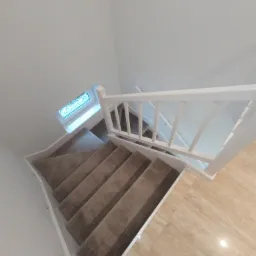 Widok z góry na schody z wykładziną w odcieniach beżu, białe ściany i balustradę, oraz marmurowe płytki na podłodze, małe okno doświetla klatkę schodową.