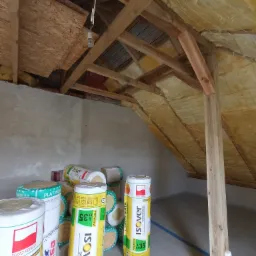 Poddasze w trakcie ocieplania, widoczne belki konstrukcyjne, zamontowana izolacja oraz rolki wełny mineralnej Isover czekające na montaż, surowy stan ścian.