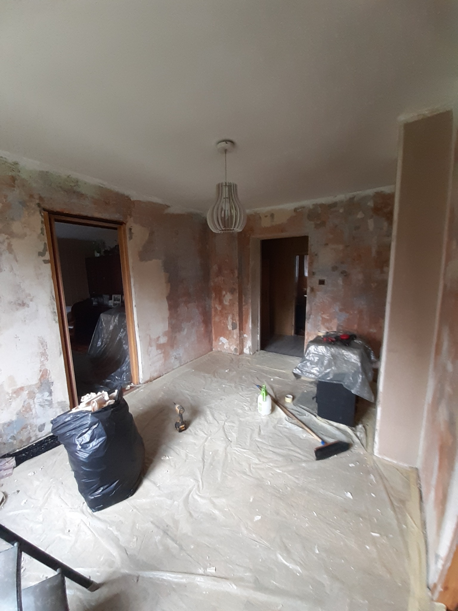 Pokój w trakcie przygotowania do prac wykończeniowych: ściany z widocznymi warstwami starej farby i fragmentami gładzi, podłoga zabezpieczona folią malarską, widoczny worek z odpadami, narzędzia...