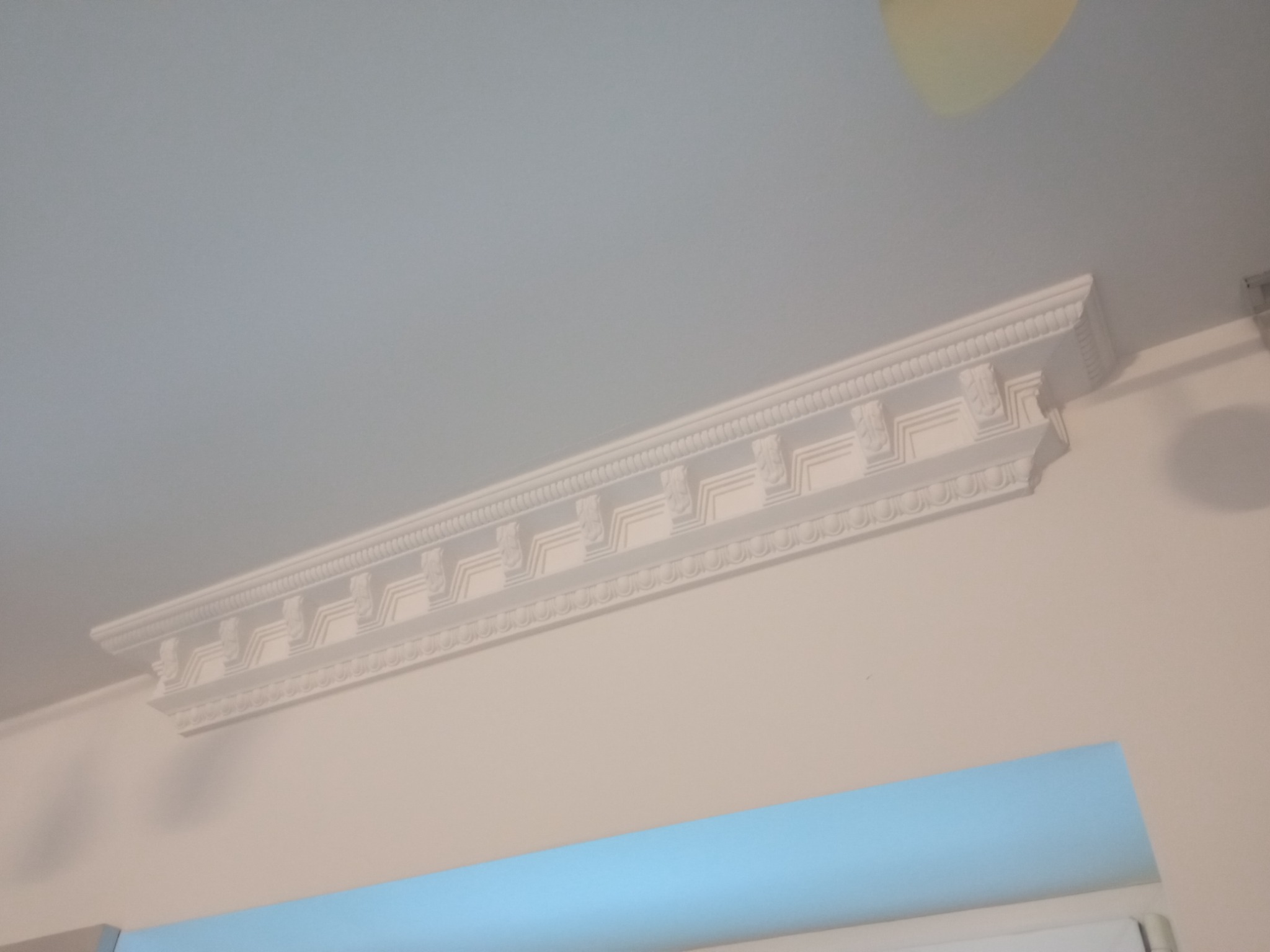 Dekoracyjny, biały gzyms sufitowy z ornamentami w klasycznym stylu, zamontowany na styku ściany i sufitu w jasnym wnętrzu.