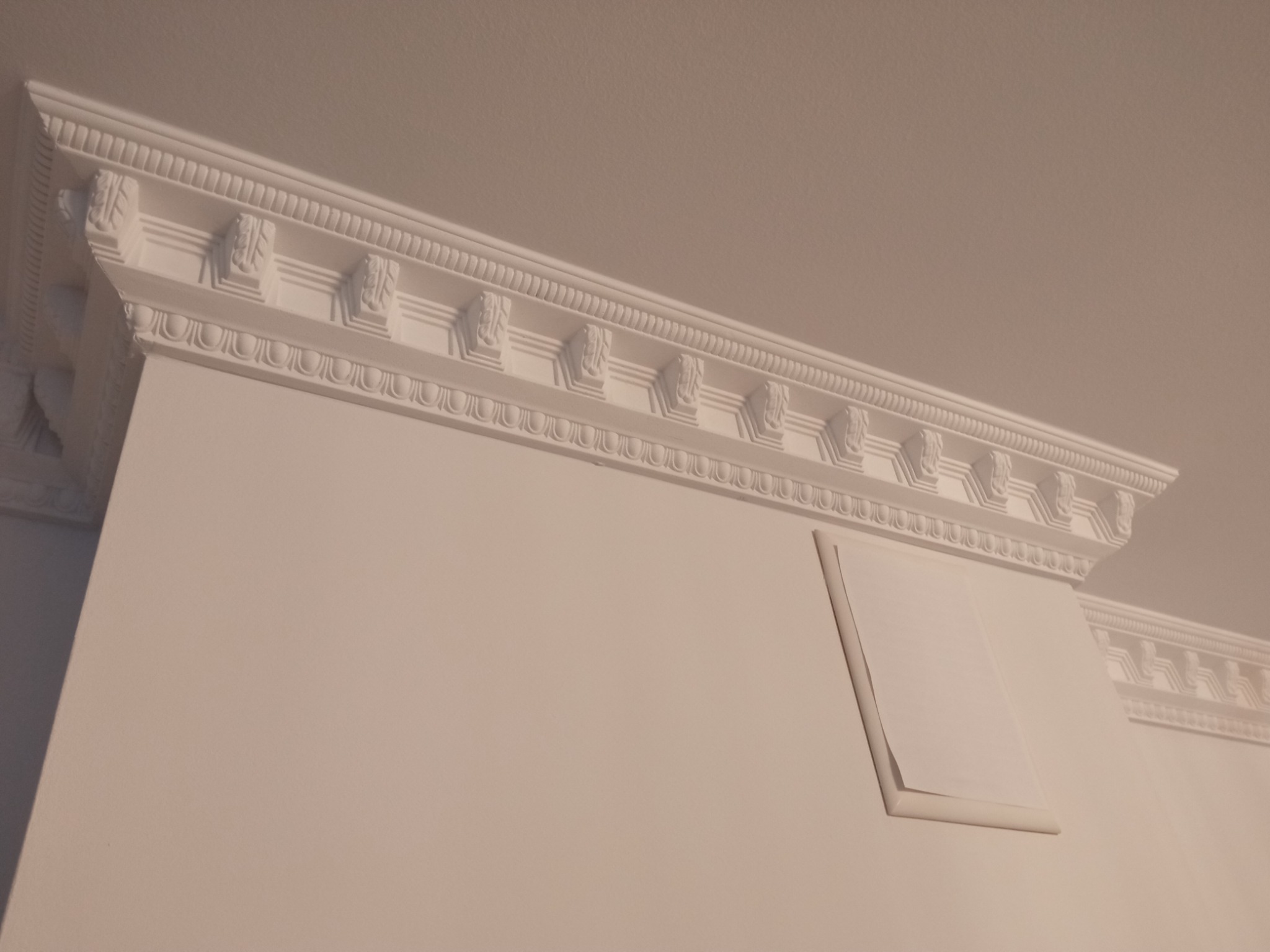 Biała ściana z ozdobnym, gipsowym wykończeniem sufitu w stylu klasycznym, widoczny fragment wentylacji.