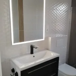 Nowoczesna łazienka z umywalką nablatową, czarną armaturą i podświetlanym lustrem, płytki ścienne z subtelnym wzorem 3D, widoczne wieszaki na przyssawki.