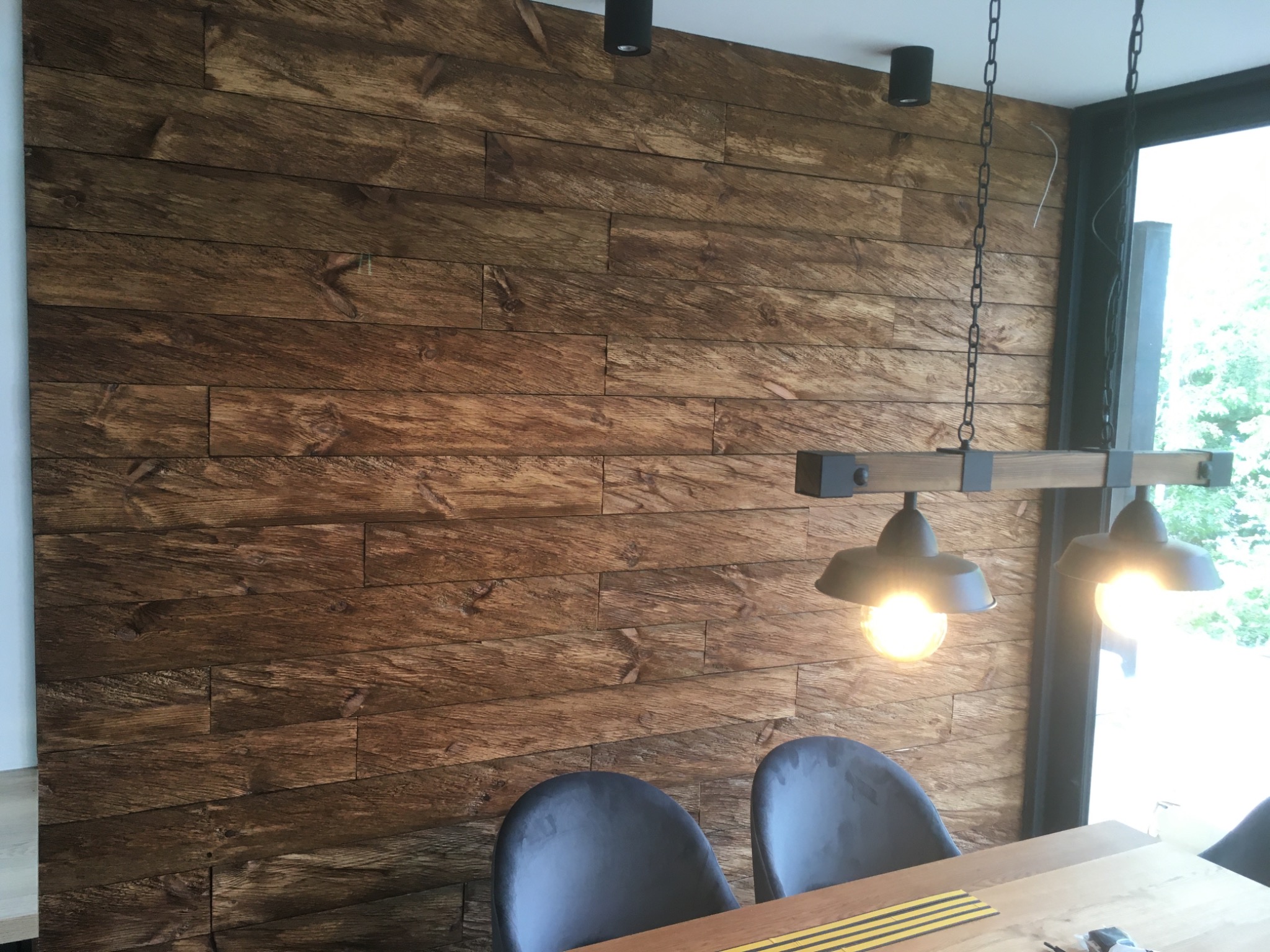 Ściana wykończona panelami z ciemnego drewna o wyraźnej fakturze, oświetlona wiszącą lampą z trzema żarówkami, widoczne krzesła i fragment stołu.