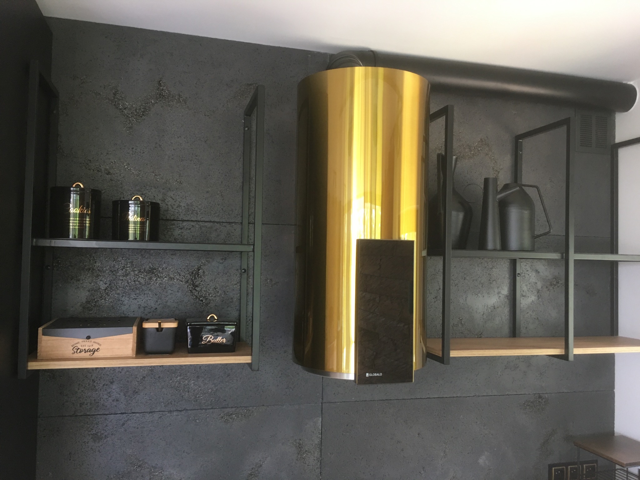 Nowoczesna kuchnia z złotym okapem cylindrycznym i czarnymi półkami w stylu loftu na tle ściany z szarego betonu architektonicznego.