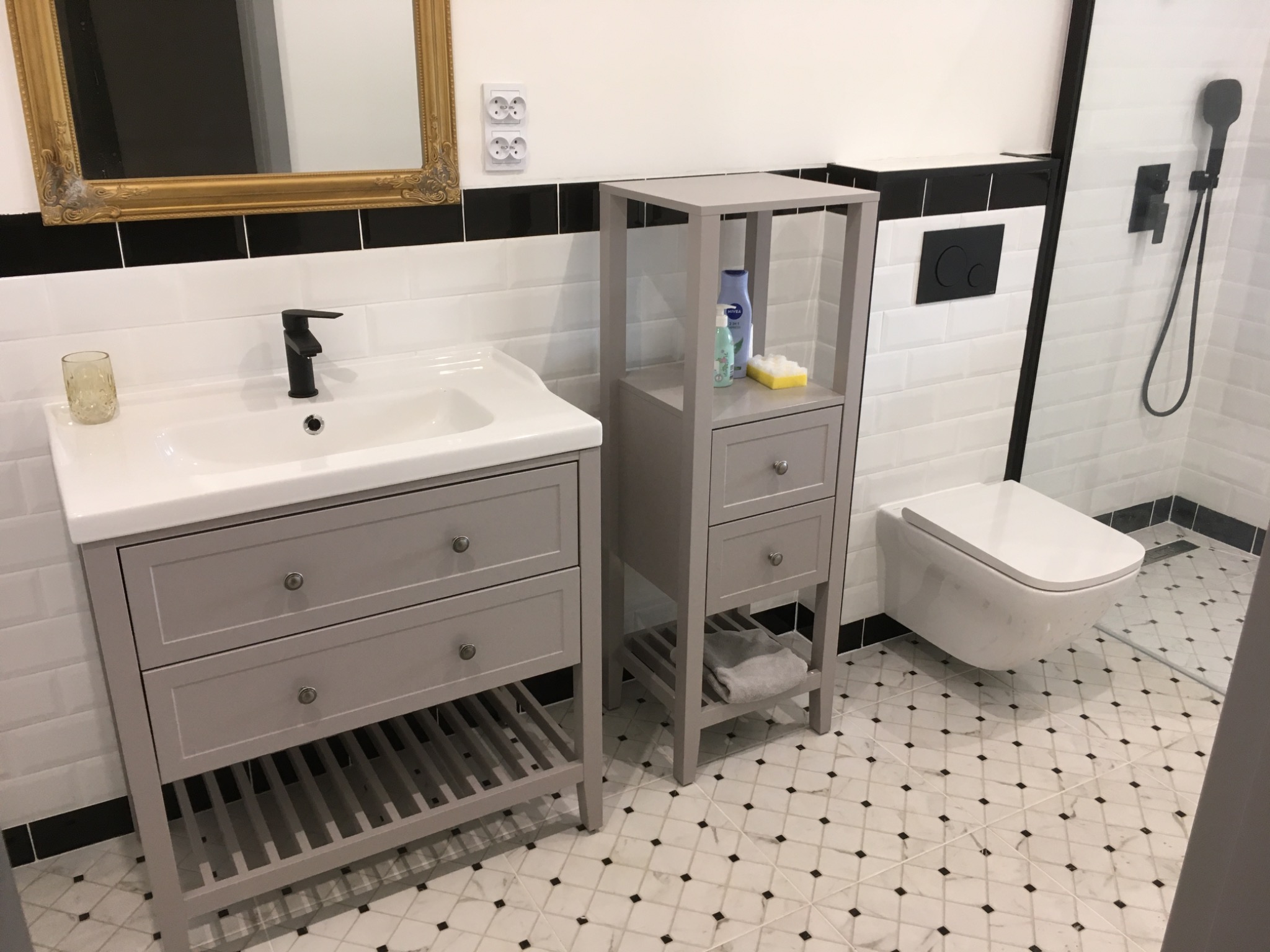 Nowoczesna łazienka z szarą szafką pod umywalką, regałem i podwieszaną toaletą. Białe płytki z czarnym paskiem i wzorzysta podłoga.