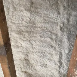 Świeżo nałożona, gęsta warstwa izolacji na poddaszu, widoczne drewniane krokwie i fragment węża aplikacyjnego z zielonym zapięciem.