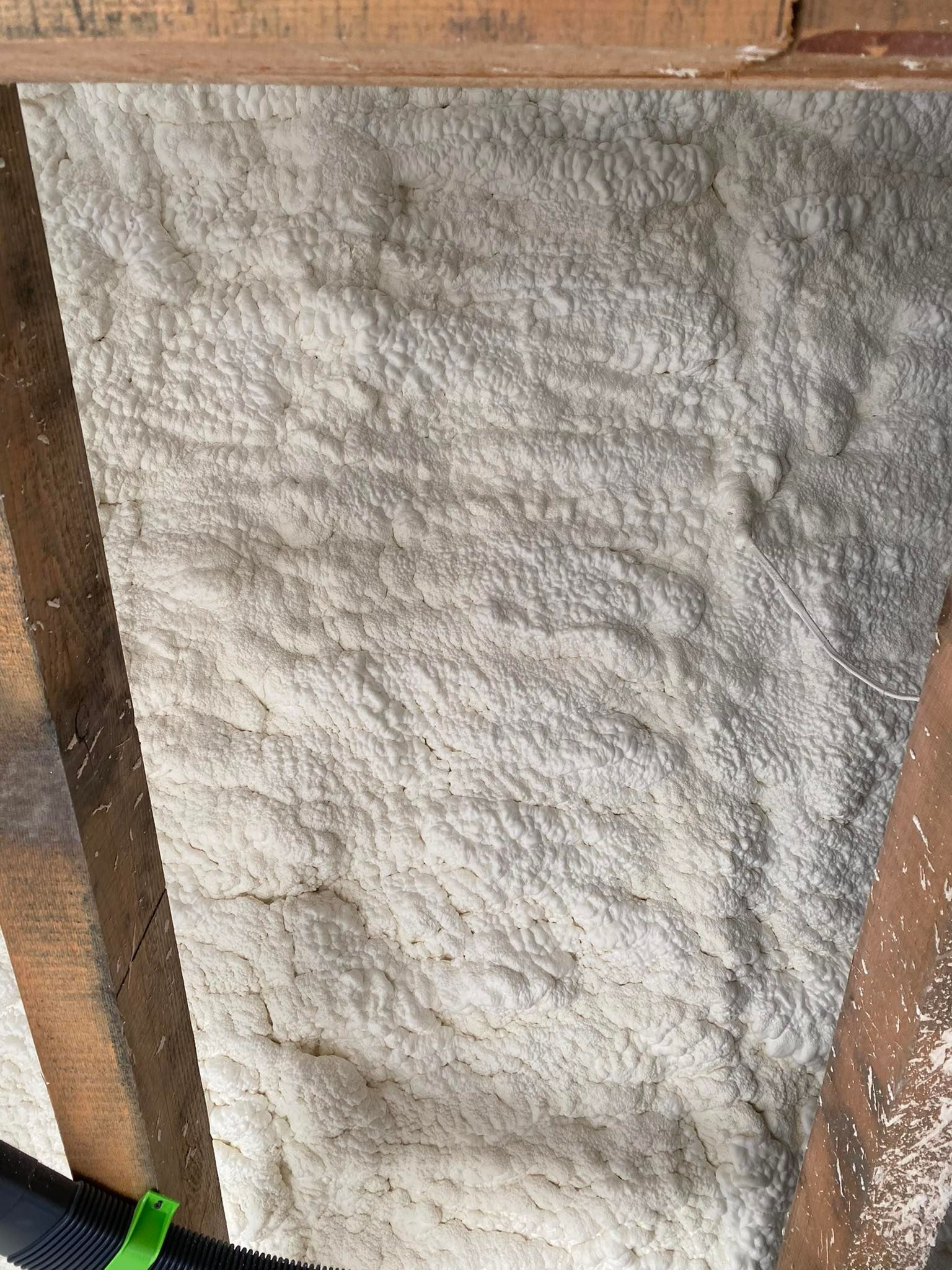 Świeżo nałożona, gęsta warstwa izolacji na poddaszu, widoczne drewniane krokwie i fragment węża aplikacyjnego z zielonym zapięciem.